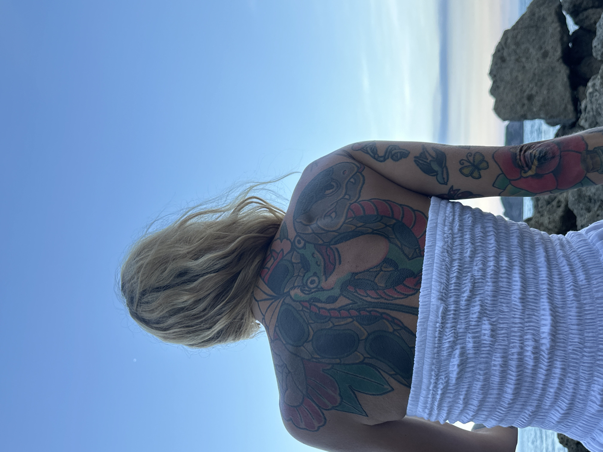 A woman with blonde hair faces away from the camera, looking out at the ocean. She has a large, colorful tattoo covering her back and part of her arm, featuring a frog and floral elements. She is wearing a white, strapless top. The background consists of a clear blue sky, the ocean, and rocky terrain. The location is identified as Mũi Né, Vietnam.