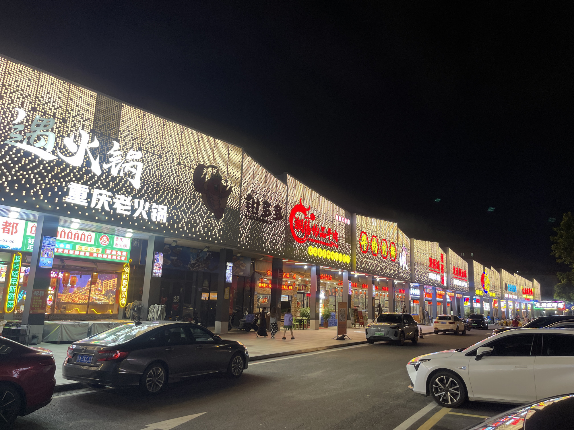 This image captures a vibrant street scene at night in Panyu District, China. The main focus is a row of restaurants, their facades illuminated by thousands of small, bright lights that create a dazzling, almost pixelated effect. Large, stylized Chinese characters are prominently displayed on the buildings, advertising "火锅" (hot pot), with "重庆老火锅" (Chongqing Old Hot Pot) being particularly visible, suggesting a culinary destination. Other signs mention "蔬菜免费" (free vegetables) and "甜品免费" (free dessert), highlighting promotions.
In front of the restaurants, a street is lined with parked cars, including a dark gray sedan with a license plate "A 3X3JO" and a white sedan. People are seen walking on the sidewalk, some appearing to be patrons entering or leaving the establishments. The overall atmosphere is lively and commercial, typical of a busy entertainment or dining area after dark. The lighting creates a festive and inviting ambiance, despite the lack of visible natural light, indicating it is nighttime. The clear sky suggests fair weather. The scene is bustling with activity, driven by the allure of dining and entertainment offered by the illuminated establishments.