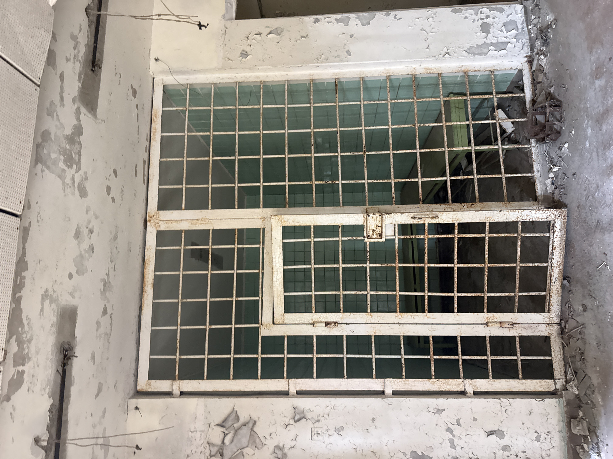 The image shows an interior view of a derelict building in Prypiat, Ukraine. A gridded window structure dominates the scene, composed of a large, fixed section and a smaller, hinged panel that appears to be partially open. The window frames and bars are painted white, but show significant signs of rust and peeling paint, indicating age and neglect. The glass within the grids is intact, but likely dirty and obscured by accumulated dust and grime. The surrounding walls are also covered in peeling white paint, exposing underlying layers and suggesting water damage. Debris is visible on the floor near the window, possibly fallen plaster or other building materials. The overall impression is one of decay and abandonment. The absence of human presence or signs of recent activity reinforce the notion of a long-abandoned space within the city.