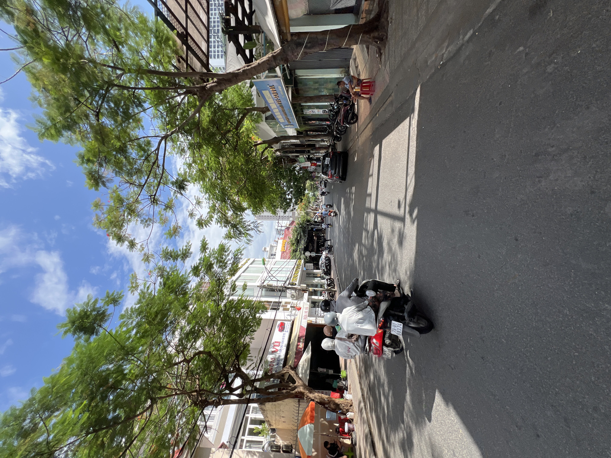 This image depicts a street scene in Nha Trang, Vietnam, characterized by a sunny day with scattered clouds. A narrow, paved road occupies the foreground, with a three-person motorcycle traveling away from the viewer. The motorcycle has a red rear panel and is driven by a person wearing a black helmet. Two passengers, one in a white hoodie and the other in a beige hoodie, are seated behind the driver. The street is flanked by buildings and trees. On the left side, buildings with light-colored facades, windows, and awnings line the street. One building displays a sign with Vietnamese text and an image of a car. A tree with lush green foliage stands prominently near the left side of the road. On the right side, a sidewalk runs alongside the buildings, with several parked motorcycles visible. A man is seated on a red stool on the sidewalk, facing the road. The buildings on the right also feature windows and awnings, and one has a blue sign with English text that appears to read "MONTESSORI PRE-SCHOOL". The overall impression is a bustling urban environment under a bright sky.