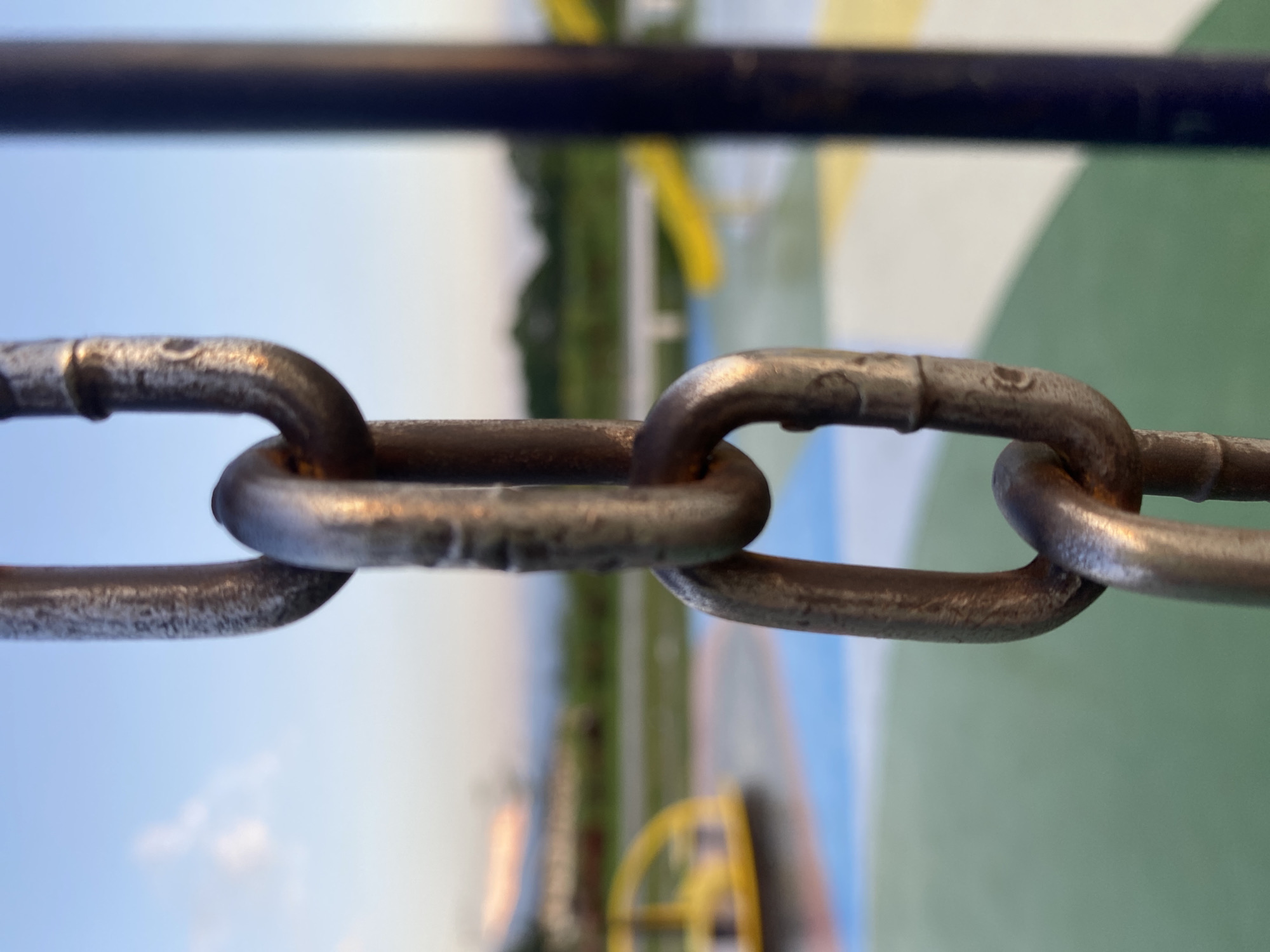 The image is a close-up, vertical shot of a metal chain, likely part of playground equipment, against a blurred background of a park. The chain links are rough and slightly rusted, indicating outdoor exposure. The background features a colorful playground surface with distinct sections of green, blue, and yellow. A yellow merry-go-round is visible on the left, and yellow slide structures are present in the distance. The sky is a soft blue with hints of sunset colors, suggesting it is late afternoon or early evening. The setting is likely a public park within Davao City, Philippines, identifiable by the common types of playground equipment and the tropical-toned sky. There are no people or animals visible in the frame.
