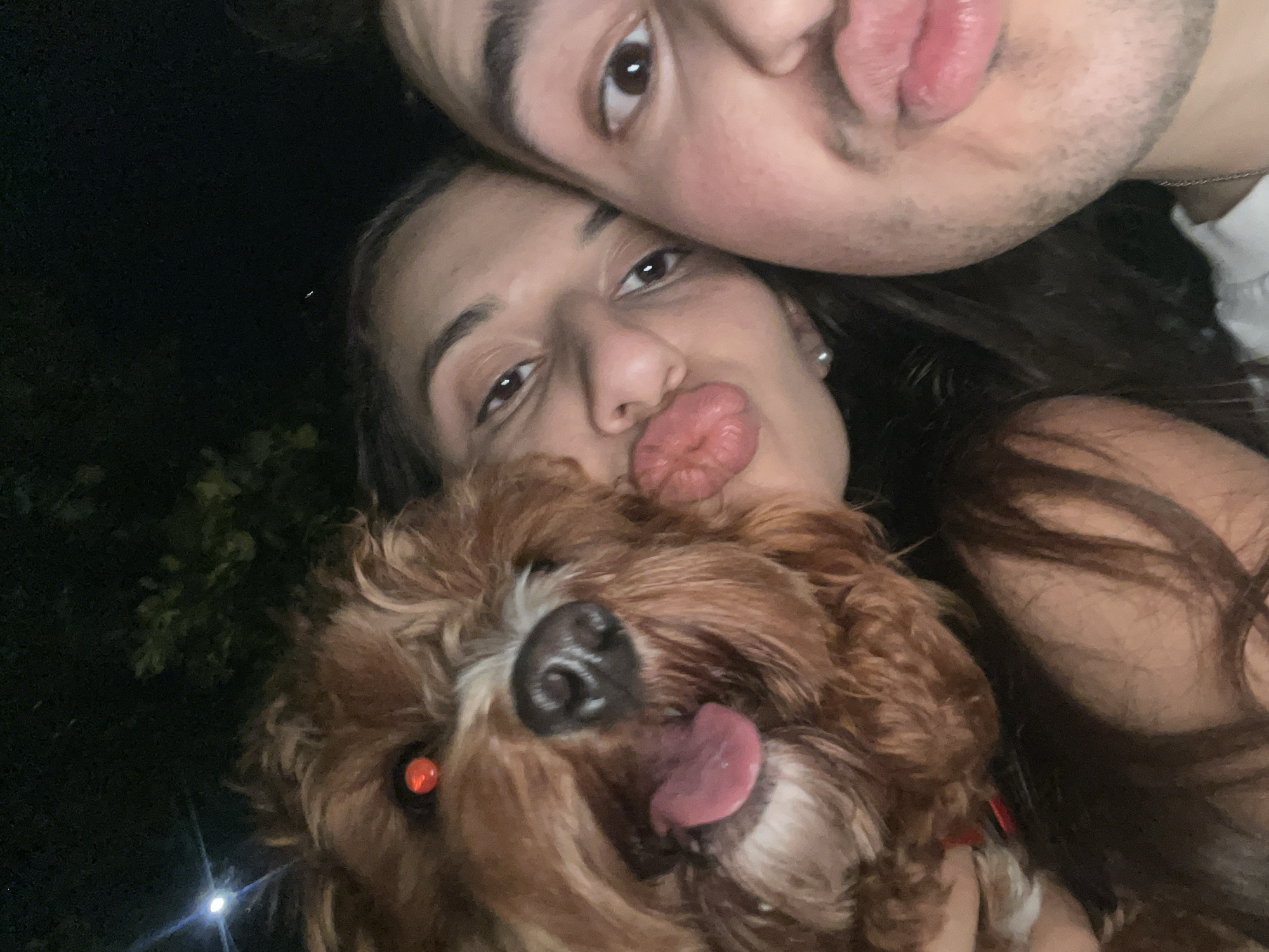A close-up, low-light selfie captures two individuals and a dog, likely taken at night due to the dark background and strong light sources creating lens flares and red-eye. The image is taken from a slightly high angle, looking down.
The subjects include a male, a female, and a medium-sized dog with brown, curly fur. The male is positioned at the top of the frame, partially visible, looking directly at the camera with dark eyes. He has dark hair, noticeable stubble on his face, and wears a silver chain around his neck over a light-colored shirt.
Below him, the female looks towards the camera with dark eyes and puckered, pouting lips, suggesting a playful expression. She has dark hair, and a pearl stud earring is visible in her ear.
The dog occupies the lower and central part of the frame, held by one of the individuals. It has its tongue playfully sticking out and a black nose. One of its eyes exhibits a distinct red-eye effect from the flash, while the other is obscured by fur. A red collar is visible around its neck.
The overall mood of the image is playful and affectionate, capturing a moment between pets and their owners. The background is mostly dark, suggesting an outdoor setting at night, with some faint glimpses of dark foliage or trees. No text is visible in the image.