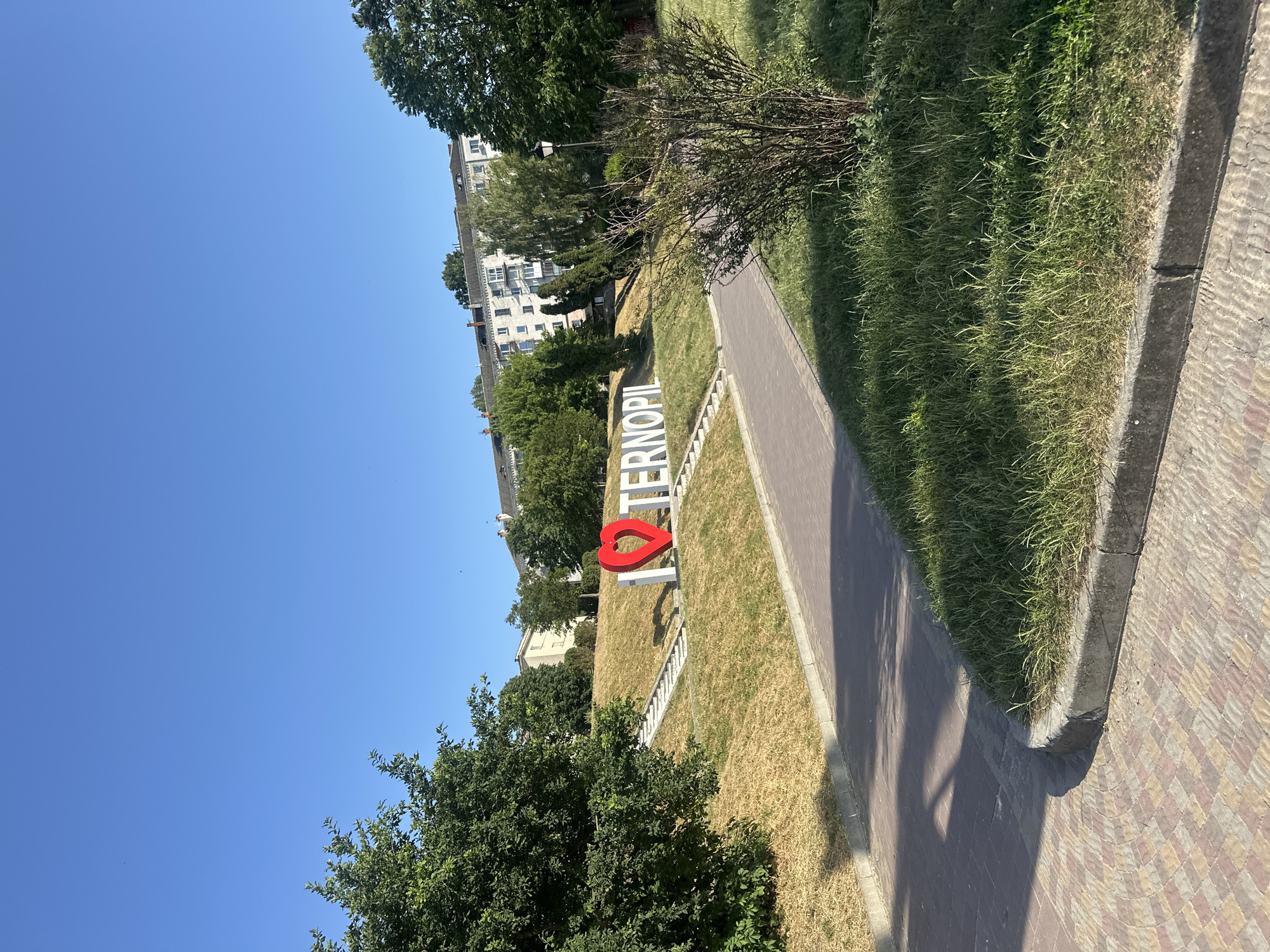 The image depicts a sunny day in Ternopil, Ukraine. The scene features a pathway made of colorful bricks that curves to the right, bordered by a strip of dry grass and a patch of lush green grass. Beyond the grassy area, a large, white sign with a red heart in the middle spells out "I LOVE TERNOPIL." The letters are bold and stand out against the backdrop of a grassy slope. Further in the background, there are several multi-story buildings, some with visible windows and balconies, suggesting an urban or suburban setting. Tall, green trees are scattered throughout the scene, with their leaves casting shadows on the ground. The sky is a clear, vibrant blue, indicating fair weather. The overall impression is one of a bright, pleasant day in a public space, possibly a park or a recreational area within the city, with the prominent sign serving as a local landmark. There are no people visible in the image. The time of day appears to be midday due to the strong sunlight and distinct shadows.