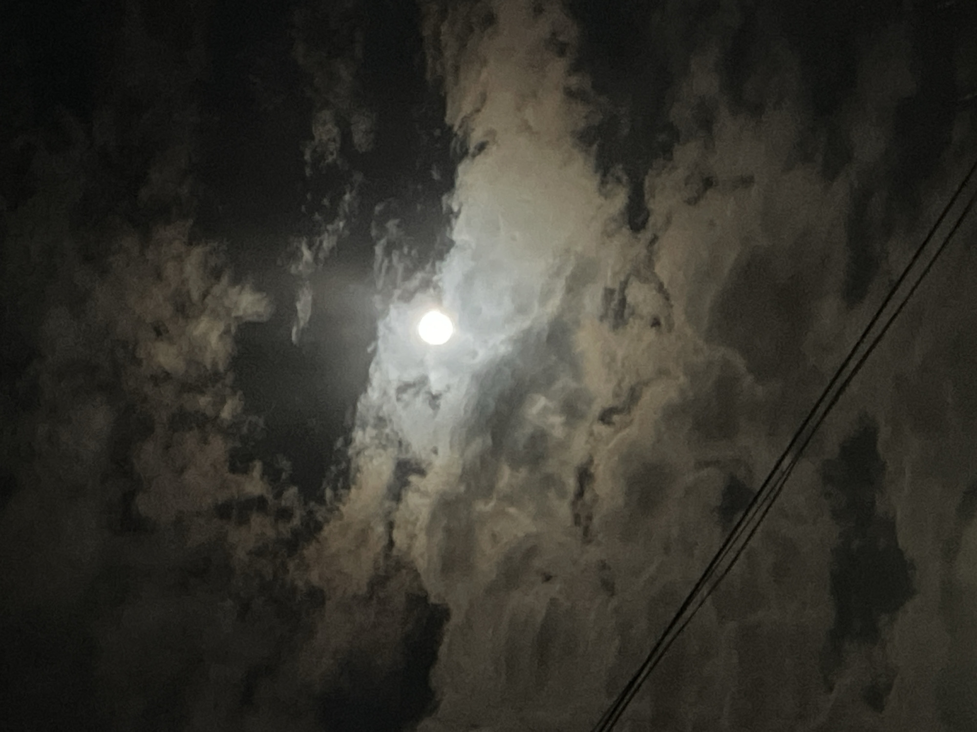 The image depicts a night scene of the sky, dominated by a bright moon shining through a layer of clouds. The clouds vary in density, with some areas appearing darker and more opaque, while others are thinner and allow more moonlight to pass through. A pair of power lines run diagonally across the right side of the frame. The location is Yerevan, Armenia. No people are visible.