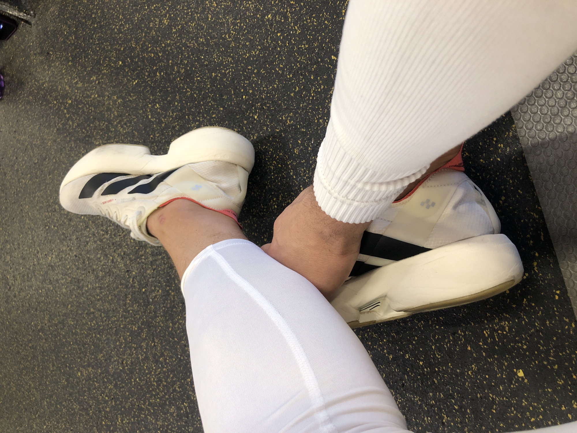 A pair of white and black Adidas athletic shoes with red accents are resting on a dark gray rubber floor. The shoes are on the feet of a person wearing white athletic pants and a white ribbed long-sleeved shirt. The scene appears to be indoors, possibly in a gym. There are no discernible landmarks or features that would indicate the location is specifically Saint Petersburg, Russia.