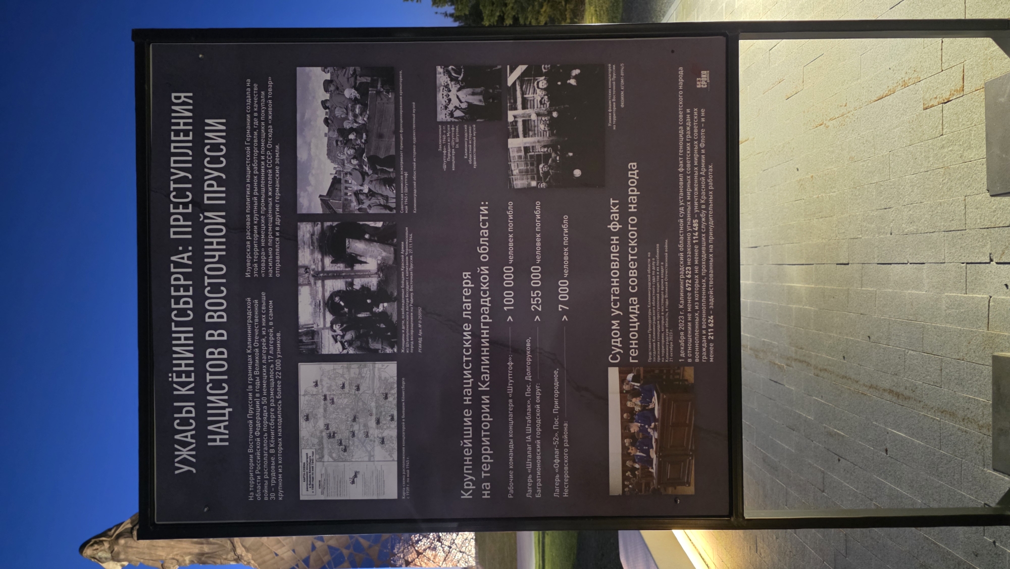 The image shows an information sign titled "Horrors of Königsberg: Crimes of the Nazis in East Prussia". The sign details atrocities committed by Nazi Germany in the East Prussia region, particularly within what is now the Kaliningrad Oblast of Russia. It mentions the presence of around 50 Nazi camps, including 17 in Königsberg, and highlights the enslavement and forced labor of Soviet citizens. The sign lists major Nazi concentration camps in the Kaliningrad Oblast and estimates the number of deaths. It states that on December 1, 2023, the Kaliningrad Regional Court established the fact of genocide against the Soviet people. The sign includes photographs of prisoners and documentation. The location of the sign is Zaitsevo (Verevskoe S/P), Russia, but the image gives no details about that location.