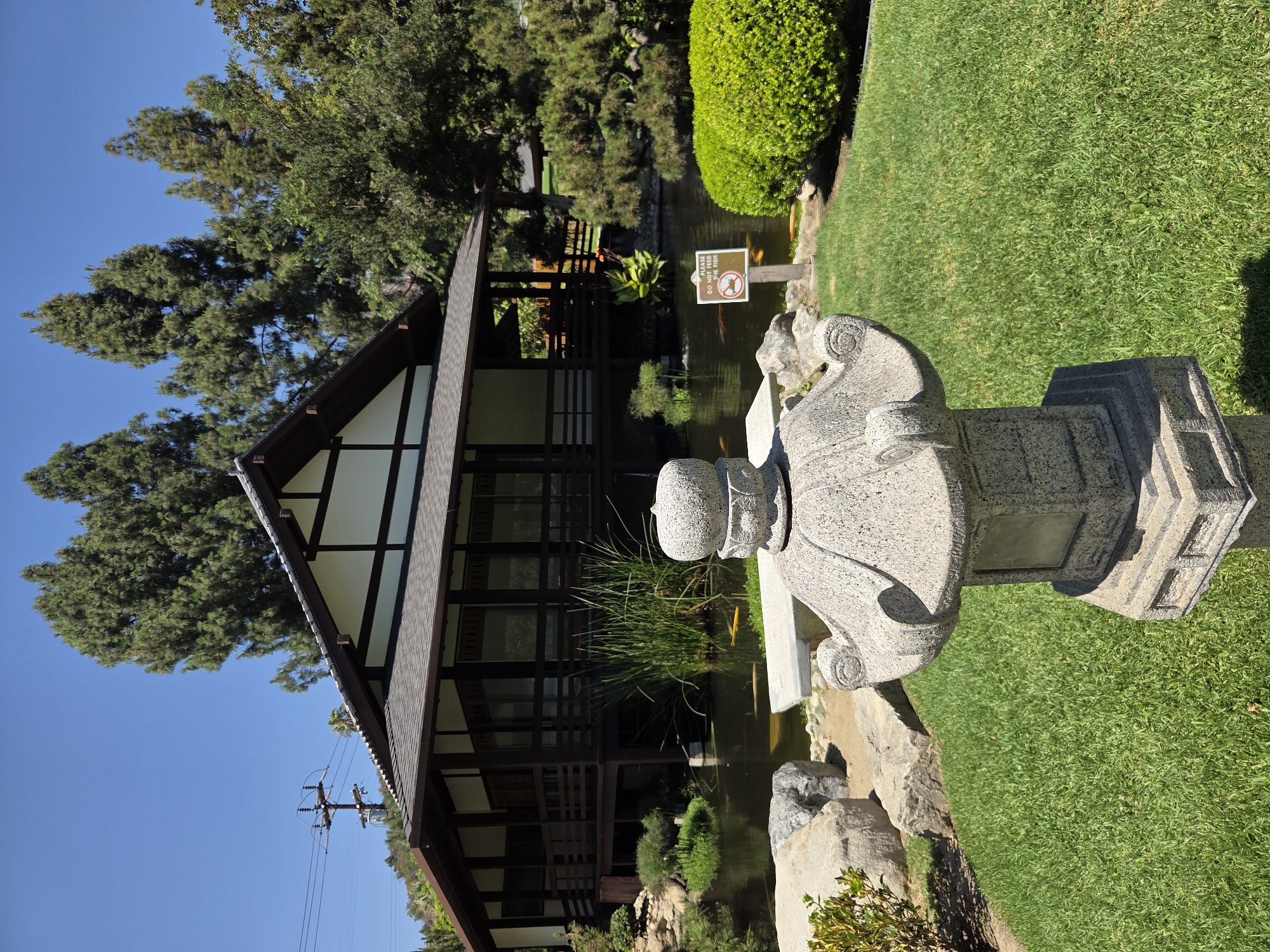 The image depicts a tranquil Japanese garden scene in Glendale, California. In the foreground, a stone lantern stands on a manicured green lawn. Behind the lantern, a calm pond reflects the clear blue sky and is populated by several orange koi fish. Tall reeds and aquatic plants grow along the edge of the pond.

Dominating the midground is a traditional Japanese-style building with dark wood beams and white walls, elevated on stilts. Verandas with railings encircle the structure, and large windows offer glimpses inside. The building is surrounded by lush trees and manicured shrubs.

A small sign near the pond reads, "PLEASE DO NOT FEED THE FISH." In the background, a hill covered with trees and a utility pole with electrical lines are visible. The overall atmosphere is serene and well-maintained, characteristic of a Japanese garden.