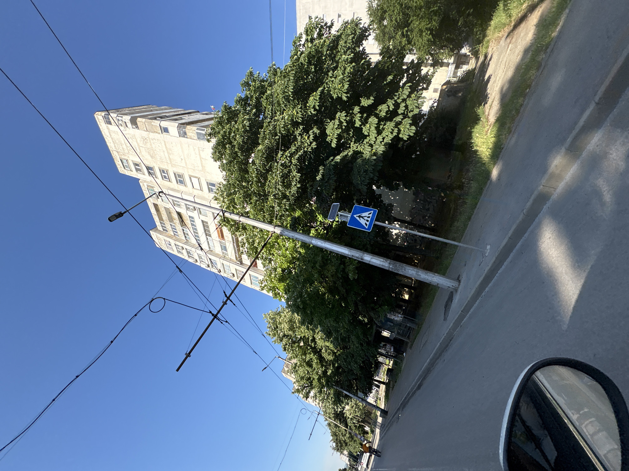In Vratsa, Bulgaria, a street scene unfolds under a clear blue sky, dominated by a tall, weathered apartment building. Trolleybus wires crisscross the sky, indicative of public transportation infrastructure. In the foreground, a car's side mirror reflects the street, suggesting the image was taken from inside a vehicle. A pedestrian crossing sign stands by the roadside, next to a pole that supports trolleybus wires and a street lamp. Lush green trees line the street, partially obscuring the apartment building and creating a sense of urban greenery. A cyclist can be seen in the distance, riding along the road. The overall environment is a typical urban streetscape with elements of both residential and transportation infrastructure.