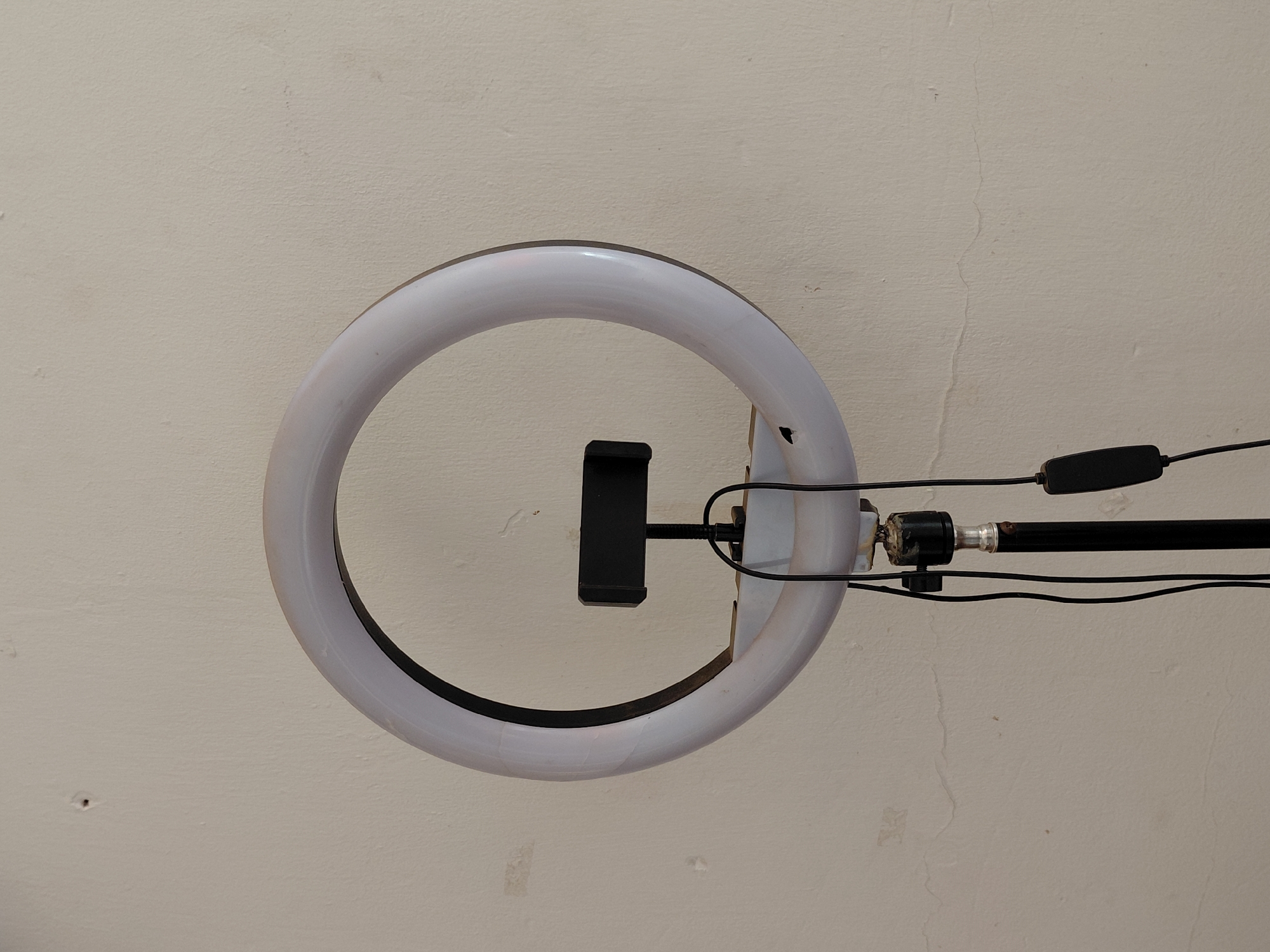The image features a white ring light suspended against a light-colored, textured wall or ceiling. The circular light has a translucent white plastic diffuser, with a darker, likely black, rim visible along its inner and outer circumference. A black smartphone holder is centrally mounted within the ring, oriented vertically.
Several black wires extend from the back of the ring light assembly. One set of wires connects to a small, black rectangular control unit, positioned towards the upper right, which appears to be a power switch or dimmer. Another black, cylindrical support arm extends horizontally from the rear of the light assembly, anchored by a metal joint.
The background is an off-white or light beige surface with a visible texture. It shows minor imperfections, including faint, hairline cracks and small, scattered marks, which are more evident in the provided close-up crops. The ring light is not brightly illuminated, suggesting it is either off or at a low setting, allowing its components to be clearly seen.
The scene depicts a static lighting setup, likely intended for photography or videography. No people are present, and no specific activity is taking place. There is no visible text within the frame. While the context states Santa Ana, Venezuela, there are no specific visual cues in the image itself to confirm this location.