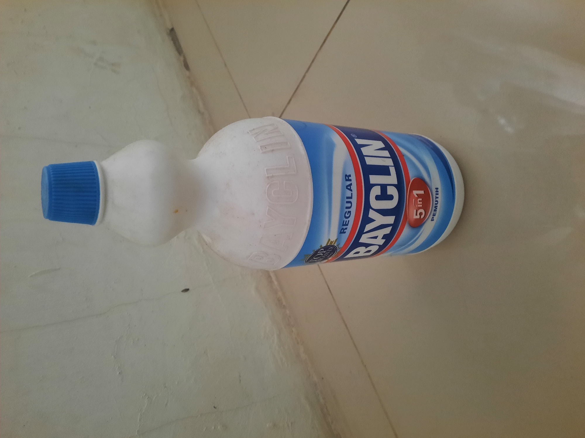 A white plastic bottle of Bayclin, a bleach product, stands on a tiled floor. The bottle has a blue ribbed cap and a label with blue and white waves. The label displays the brand name "BAYCLIN" in large white letters, with "REGULAR" printed above it. Below the brand name, a red circle with "5 in 1" is visible, and beneath that, the word "PEMUTIH" is written. The bottle is positioned in front of a textured, light-colored wall. The floor is tiled with light-colored square tiles. The scene appears to be indoors, possibly in a domestic setting, but no specific location in Lhokseumawe, Indonesia, can be determined from the image alone.