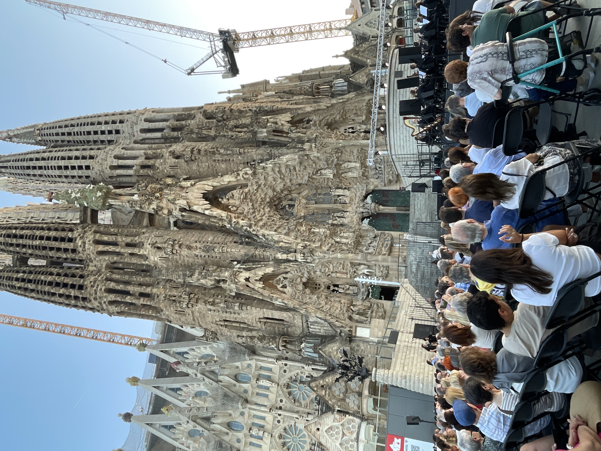 This image depicts a gathering of people seated in chairs, facing a grand, unfinished stone structure. The architecture is highly intricate and ornate, with numerous towers, spires, and detailed carvings. Two large construction cranes are visible in the background, one orange and one yellow, suggesting ongoing construction or renovation. The sky is clear and blue, indicating fair weather. The scene appears to be outdoors, possibly an assembly area in front of a significant monument or cathedral.
The people are seated, some looking towards the structure, while others are engaged in conversation or looking at their phones. Their attire is casual, suggesting a daytime event. The atmosphere seems relaxed and observational. The lighting suggests it is daytime, likely afternoon given the light angle on the structure and the people. The architectural style strongly suggests the Sagrada Familia in Barcelona, Spain, given its distinctive, organic, and unfinished appearance, and the presence of cranes.
Visible text is minimal and difficult to decipher clearly, but one sign appears to have red and white elements and possibly stylized lettering, common for signage at tourist attractions or event venues. The overall impression is of a group of visitors or attendees observing a remarkable architectural site during daylight hours in an urban setting known for its historical and artistic landmarks.