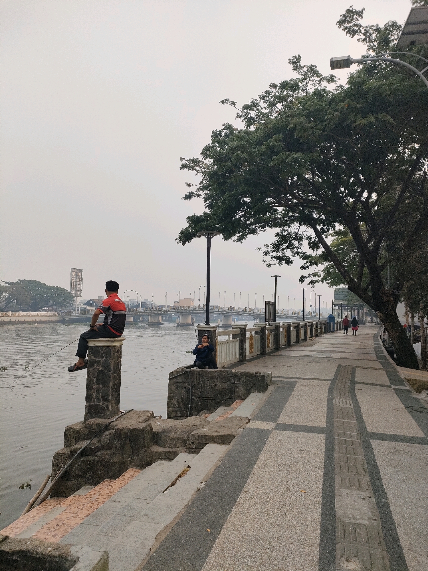 The image depicts a riverside promenade in Banjarmasin, Indonesia, likely along the Martapura River. In the foreground, a man wearing a red and gray shirt and dark pants is seated on a stone pillar, fishing with a rod and line. To his right, another person in dark clothing sits on a lower stone structure. The promenade features a patterned walkway with alternating light and dark gray sections, bordered by a white railing. A large tree with dense green foliage overhangs the walkway on the right side, with a street lamp visible near its trunk. In the background, a bridge spans the river, with several street lamps lining the opposite bank, suggesting an urban setting. The sky is overcast, contributing to a muted, hazy atmosphere.