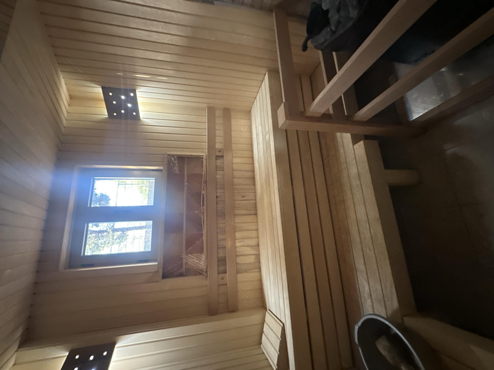 The image shows the interior of a wooden sauna. The walls and ceiling are constructed of light-colored wooden planks. There are two levels of wooden benches, with the upper bench being narrower. A small window with two panes is visible above the upper bench, offering a view of trees and a bright sky. Two square wall-mounted lights with multiple small bulbs are present, one on the left and one on the right side of the room. A small wooden stool is placed on the lower bench on the left side of the image. A metal bucket with a wooden handle is on the floor to the left. In the foreground on the right, part of a wooden structure, possibly another bench or a support, is visible, along with a black object that appears to be a sauna heater with stones and a ladle. The overall impression is of a clean and functional sauna space. The location is Brca, Montenegro.