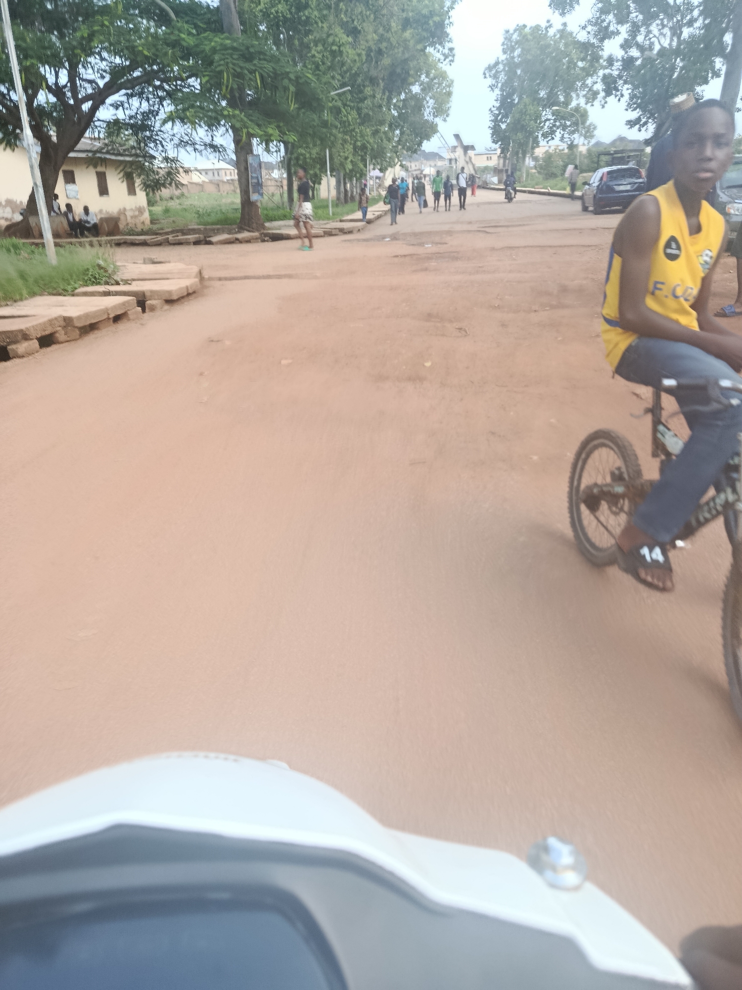 The image captures a street scene in Rafin Par, Nigeria, likely on a dry, dusty day. In the foreground, a white scooter or motorcycle is partially visible, indicating the photographer's perspective. The main subject is a young person on a bicycle, wearing a yellow sleeveless shirt with blue trim and dark jeans. They have dark hair and are looking towards the camera. The bicycle has a number "14" visible on the left sandal of the rider. In the background, several other people are walking along the dirt road. Some are dressed casually, while others appear to be in athletic wear. Trees with green foliage line the sides of the road, and buildings, possibly homes or shops, are visible in the distance. A dark-colored car is parked on the right side of the road. The overall environment suggests a casual, outdoor gathering or a typical day in the community.