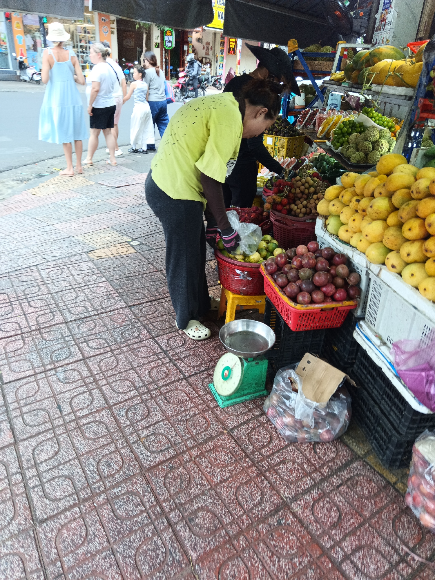 The image depicts a street scene in Nha Trang, Vietnam, likely a market stall. In the foreground, a person in a yellow shirt and dark pants is using a green mechanical scale to weigh fruit. Several baskets of fruit, including mangoes, passion fruit, and rambutan, are visible. Behind the person, other individuals are seen browsing or working at the stall. The background shows a street with buildings and a motorbike. The ground is paved with decorative tiles.