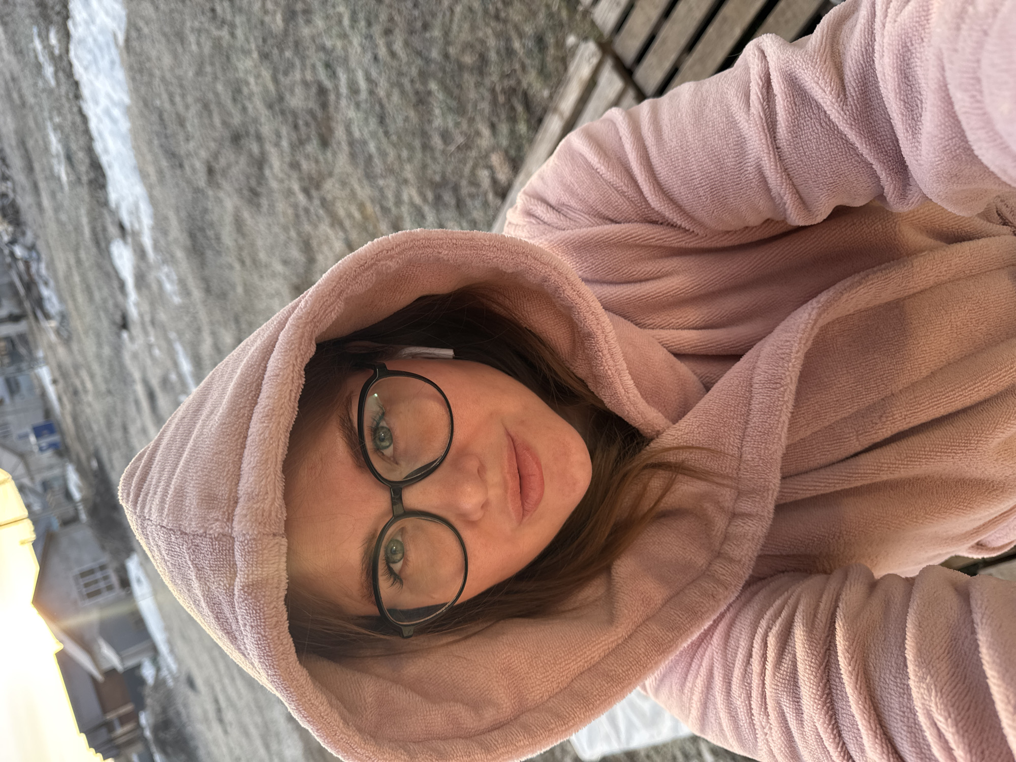 A woman with glasses and a pink hooded sweatshirt is looking at the camera. The location appears to be outdoors in Ферма, Russia, judging by the snow-covered ground and the architecture of the nearby buildings. The woman is likely on a porch or balcony.