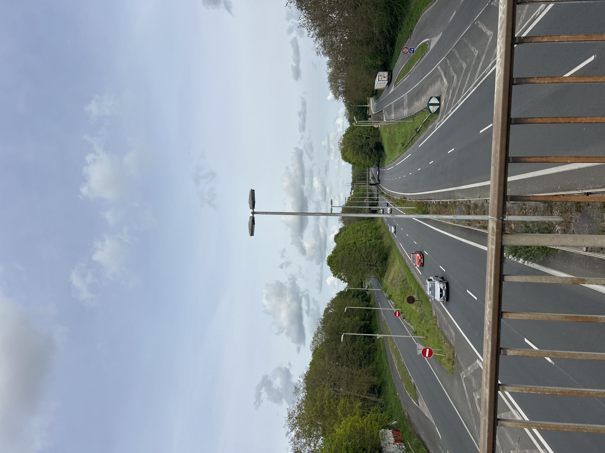 A vertical shot from a bridge looking down at a highway. Two three-lane highways run parallel to each other, separated by a grassy median with trees. Street lights line the median and the outside of each highway. Vehicles are visible on both highways, including cars and a truck. The sky is overcast with clouds. A rusted metal railing is visible in the foreground of the bridge. The highway is in Noisiel, France.