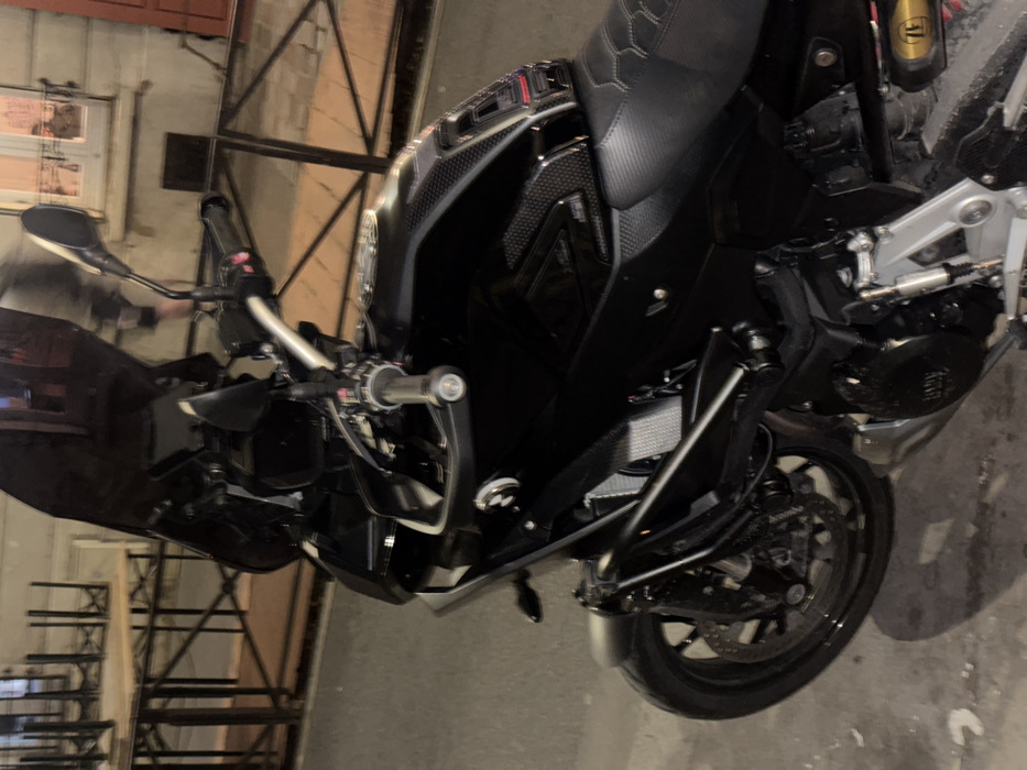 A black motorcycle dominates the frame, angled to display its front and side. The seat features a textured pattern. Handlebars, mirrors, and a front wheel with Brembo brakes are visible. The engine and surrounding mechanics are shown in detail. A portion of the background features a window and metal framework. The ground around the motorcycle appears to be concrete.