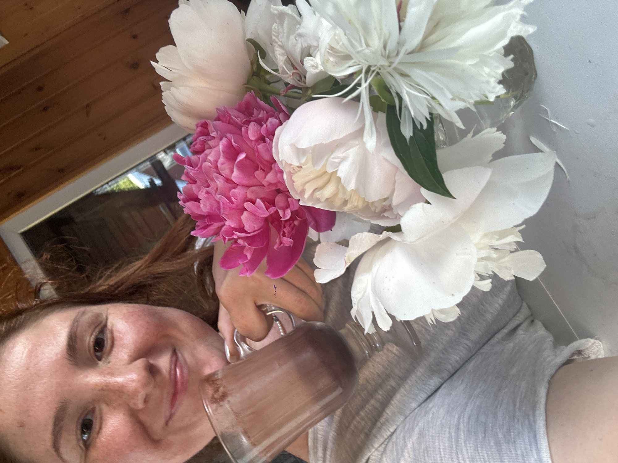 A close-up selfie shows a woman holding a clear glass mug of chocolate drink in her left hand. She is smiling and looking towards the camera. In her right hand, she is holding a bouquet of peonies. The flowers are a mix of bright pink and white. The background is blurred, but appears to be an indoor setting with wooden walls. The overall lighting suggests it is daytime. There is no identifiable information in the image that would place the location in Nizhny Novgorod, Russia.