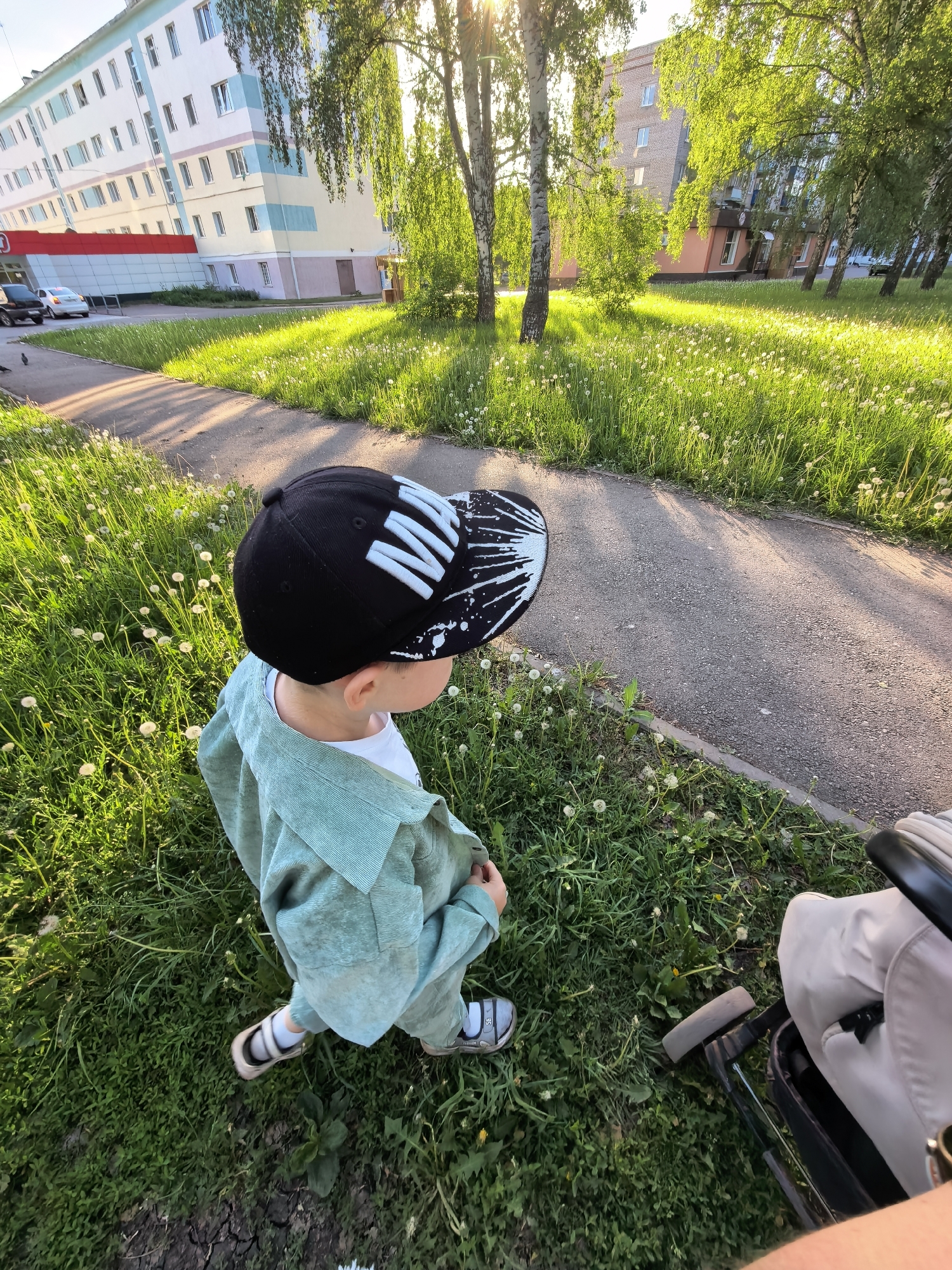 A young child stands on a grassy lawn, partially covered with dandelions, in an outdoor setting. The child is wearing a black cap with white markings, a light green outfit, and sandals. A stroller is visible on the right side of the image. In the background, there is a paved path and several buildings. The image was taken in Sterlitamak, Russia. The sun is shining brightly, casting shadows on the ground.