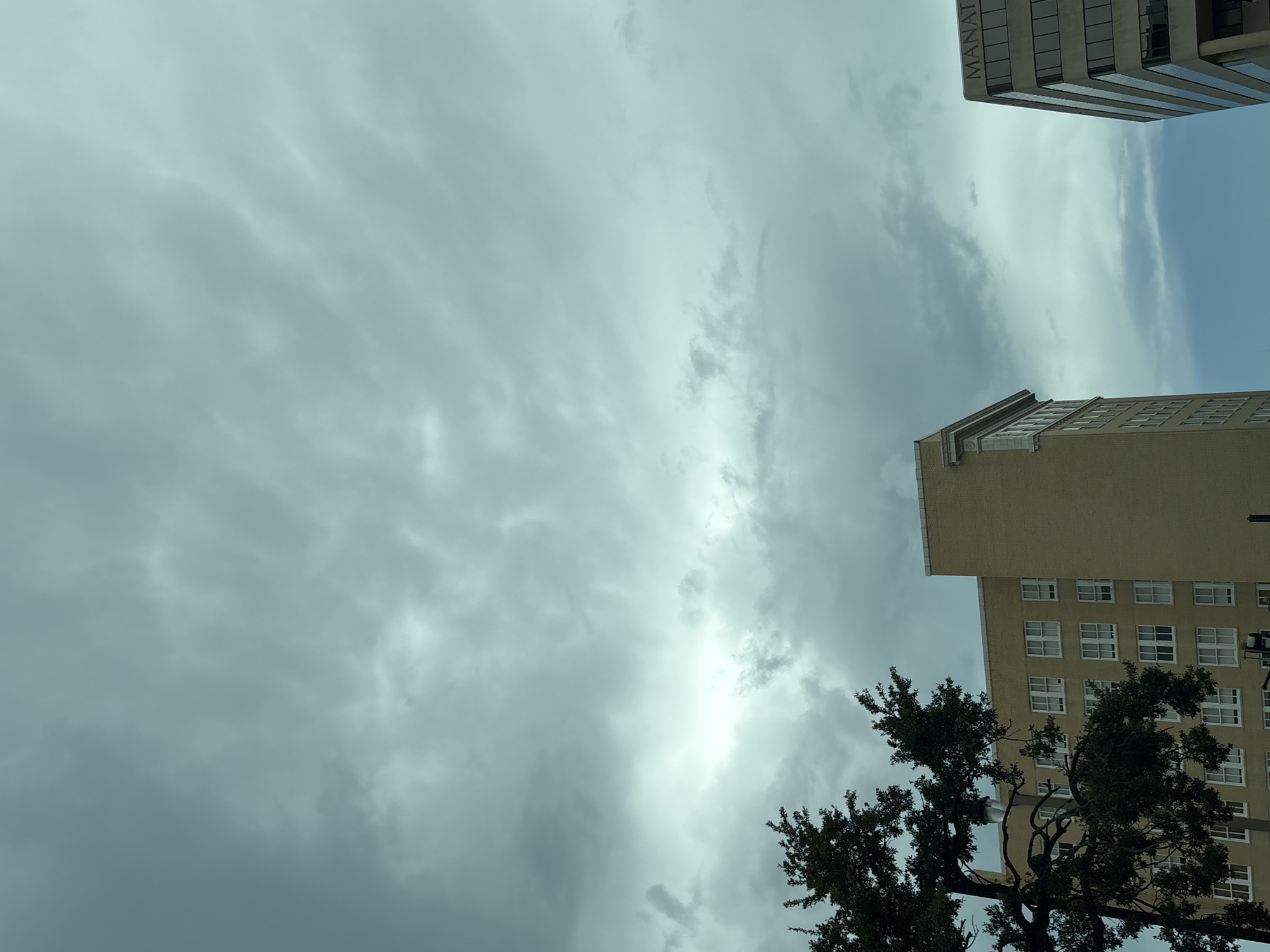 The image depicts an overcast sky over the city of Bradenton, Florida, USA. In the foreground on the left, a large tree with dark green foliage partially obscures a light yellow brick building. This building has numerous white-framed windows arranged in a grid pattern. To the right, a modern glass-fronted building is visible, with the partial word "MANAT" in dark lettering across its upper facade. The sky is filled with gray and white clouds, suggesting a cloudy or potentially stormy day. The scene appears to be an urban environment, likely a downtown area, given the presence of the buildings. No people are visible in the frame.