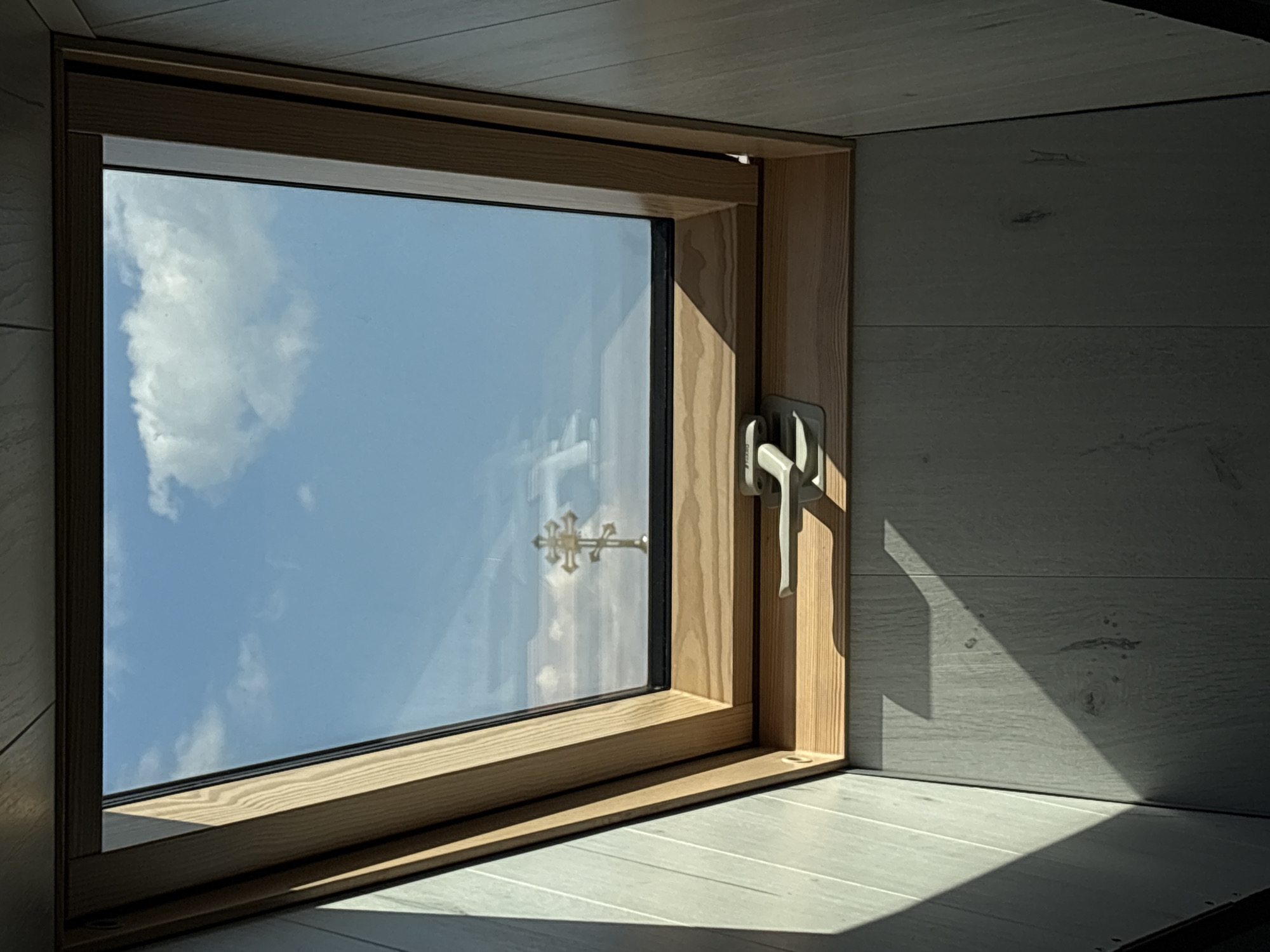 The image shows a skylight in what appears to be an attic or an upper floor room. The skylight has a wooden frame and a metal latch mechanism. Through the glass of the skylight, a clear blue sky with some white clouds is visible. In the sky, a reflection of a golden Orthodox cross is visible. The walls of the room are lined with light-colored wooden panels, and sunlight casts distinct shadows on the walls. The scene is viewed from below, looking up into the skylight. There is no indication of the city of Bobrov, Russia, in the image itself.