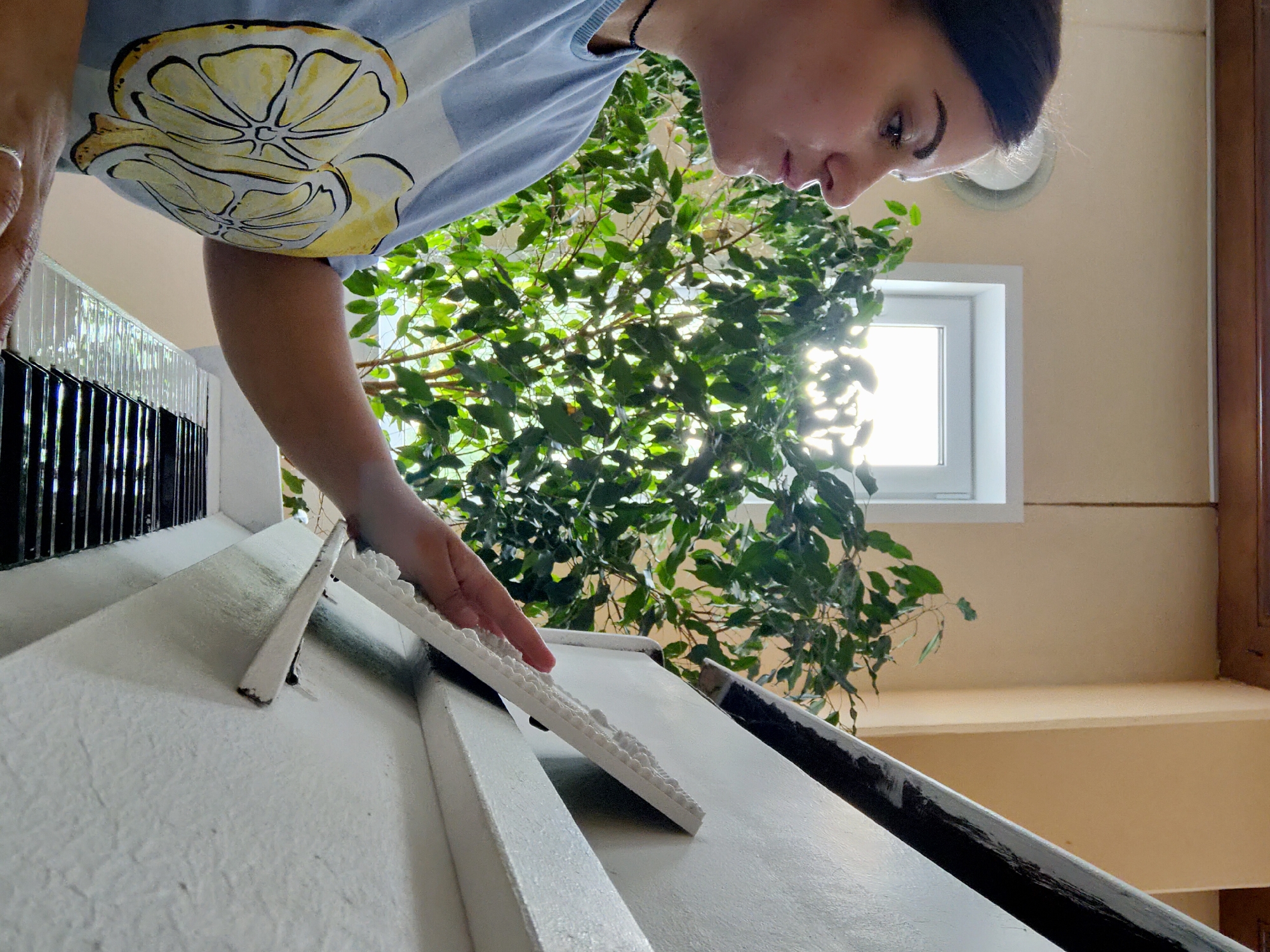 A high-angle, Dutch tilt photograph captures a person engrossed in an indoor task. The main subject is a person, likely female, seen from the chest up and leaning forward, occupying the upper left quadrant of the frame. They have dark hair tied back and wear a light blue short-sleeved t-shirt featuring a prominent yellow lemon slice design. A silver ring is visible on their left hand. Their face is in profile, showing a focused expression as they look down at the work in front of them.
The activity involves handling white decorative trim. The person's right hand holds a piece of textured white trim, aligning it with another piece already positioned on a broad white surface, which appears to be a banister or railing. This suggests they are either installing, repairing, or painting these decorative elements. Below the main white surface, a dark, possibly black, section of the railing is visible.
The scene is set indoors, with natural light streaming through a white-framed window in the background, indicating it is daytime. A large, leafy green plant, possibly a ficus, fills a significant portion of the background, positioned between the person and the window. The walls are a warm, light beige or cream color. The setting could be a hallway, staircase, or large interior space in Марковский, Russia. No visible text is present in the image. The overall impression is one of concentration and meticulous work.