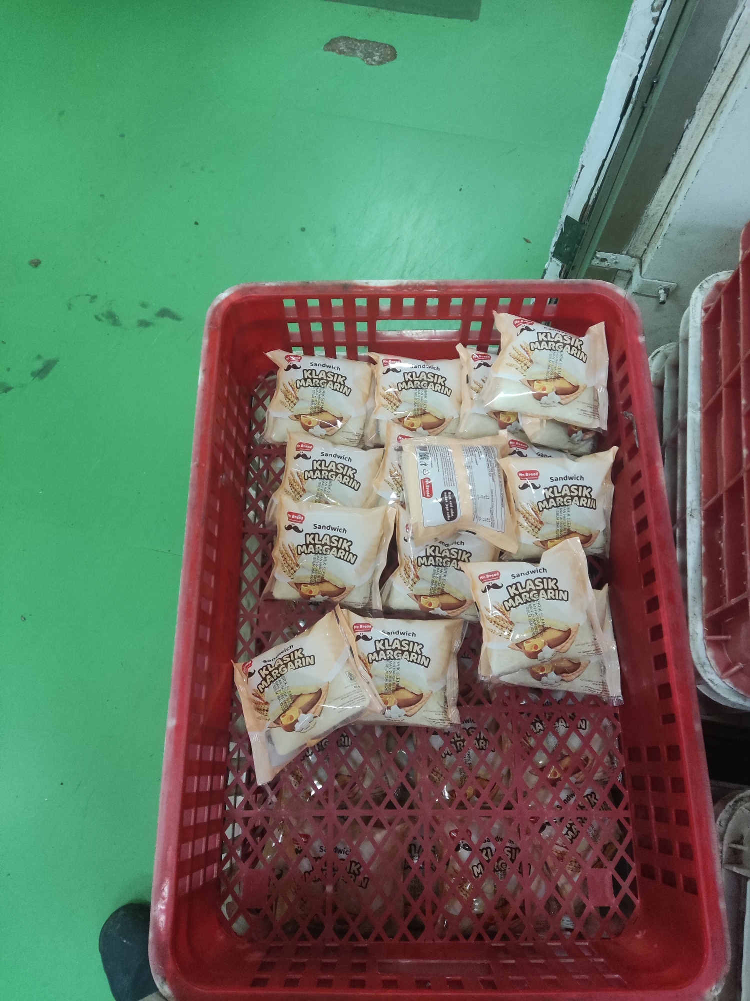 In Trihanggo, Indonesia, a red plastic basket filled with individually wrapped sandwiches is placed on a green floor. The sandwiches are labeled "Sandwich Klasik Margarin" and feature a cartoon illustration of a sandwich with margarine and a glass of milk. Some of the sandwiches are stacked in multiple layers within the basket, while others are more loosely arranged. The green floor appears to be a concrete surface, possibly in a warehouse or production facility. The overall lighting in the scene is bright, suggesting it is daytime.