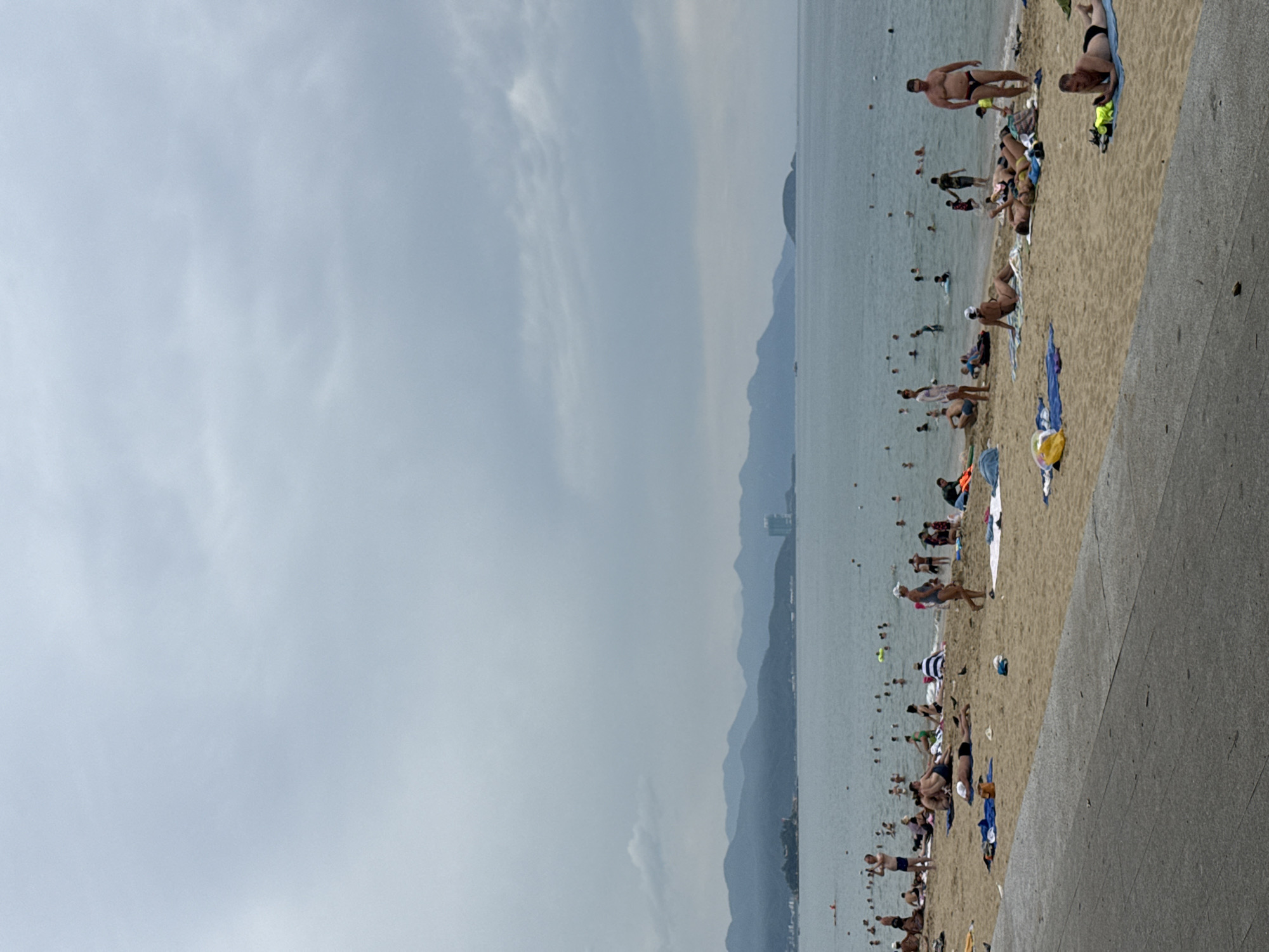The image depicts a crowded beach in Nha Trang, Vietnam, on a cloudy day. The beach is sandy and slopes down to the calm, gray water. Numerous people are scattered across the beach, some sunbathing on towels and mats, others swimming or wading in the sea. There are also people walking along the shore. The water is populated with many individuals enjoying the ocean. In the background, a range of green mountains forms the coastline, with a tall, modern building visible on the right side of the mountain range, indicating an urban presence near the natural landscape. The sky is overcast with heavy clouds, contributing to the muted lighting of the scene.