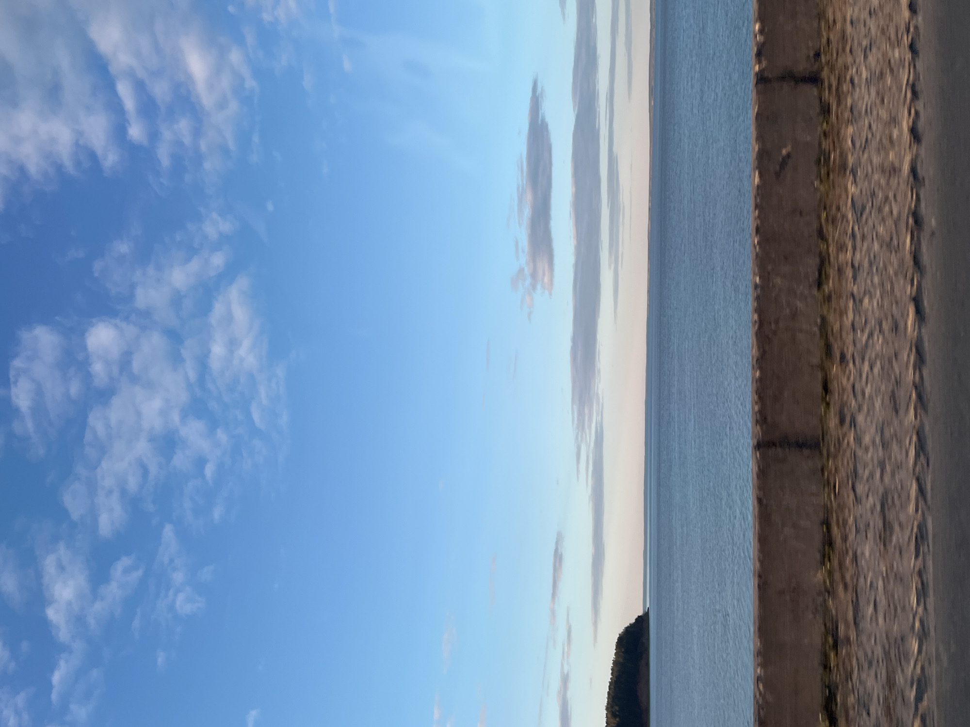 The image presents an outdoor scene with the sky occupying a significant portion, filled with light blue sky. Towards the right, a body of water stretches towards the horizon. The rightmost portion of the image shows the edge of what appears to be a paved area or sidewalk, followed by a dark-colored surface like asphalt. The location is in Noviy, Russia.