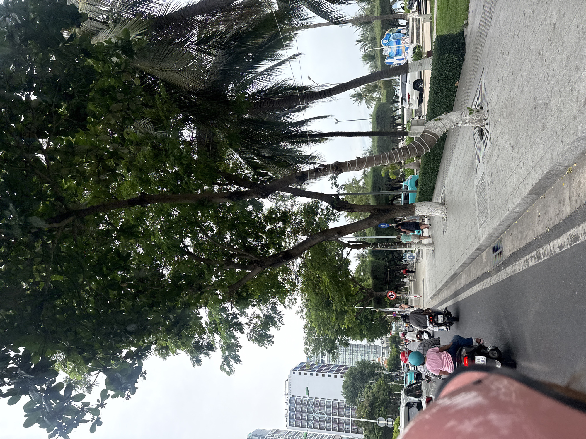 This image captures a street scene in Nha Trang, Vietnam, likely along a coastal boulevard given the presence of palm trees and a building that appears to be a hotel. The foreground is dominated by the back of a pink helmet, suggesting the photo is taken from the perspective of someone riding a motorcycle. In the street, several motorcycles are in motion. One motorcycle in the middle distance carries two people: a driver in a gray jacket and a passenger wearing a pink shirt and a bright blue helmet. Another motorcycle to the left has a driver wearing a red helmet. In the background, modern high-rise buildings, including one with "NOVOTEL" signage, line the street, indicating an urban environment. A sidewalk runs alongside the street, lined with trees and palm trees. People are seen walking on the sidewalk. The sky is overcast. A white SUV is parked on the right side of the street near a decorative display featuring a blue wave motif. The overall scene depicts a typical day in a bustling Vietnamese coastal city.