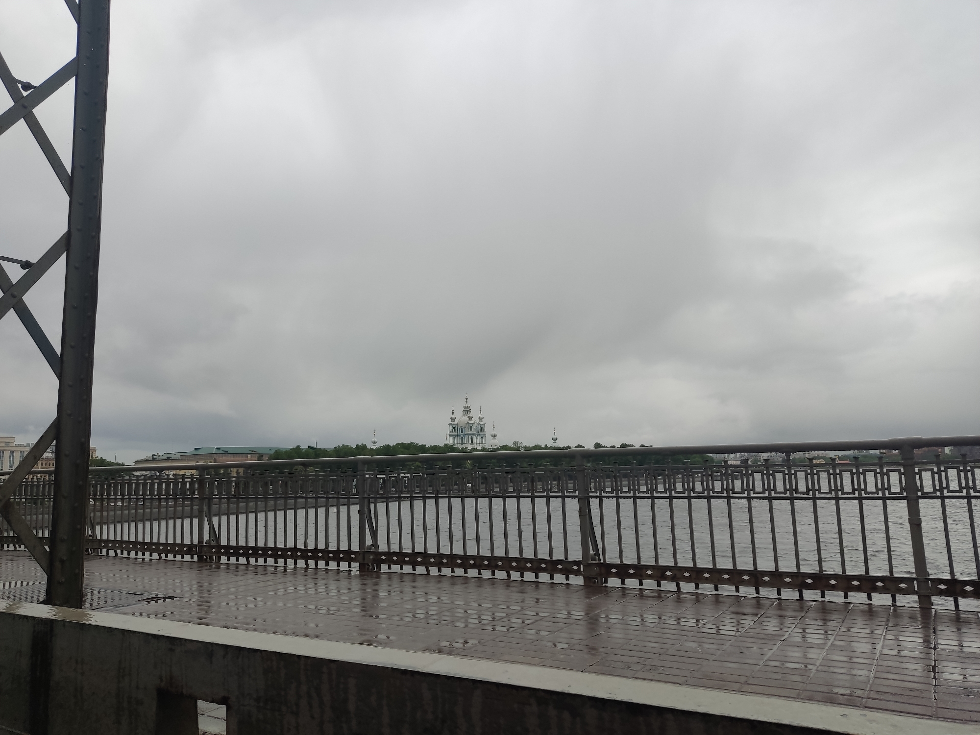The scene is an outdoor view on a cloudy day in Saint Petersburg, Russia. The vantage point is from a bridge overlooking a body of water, likely a river or canal, with a metal railing separating the viewer from the water. The bridge's surface is wet, suggesting recent rain.
In the distance, a distinctive building with multiple domes and spires is visible, which identifies it as the Smolny Cathedral, a major landmark in Saint Petersburg.
To the left of the frame, there is a metal structure, possibly part of the bridge's framework. A concrete barrier is at the bottom of the frame. The overcast sky dominates the upper portion of the image. The scene is devoid of people, emphasizing a quiet, somber atmosphere.