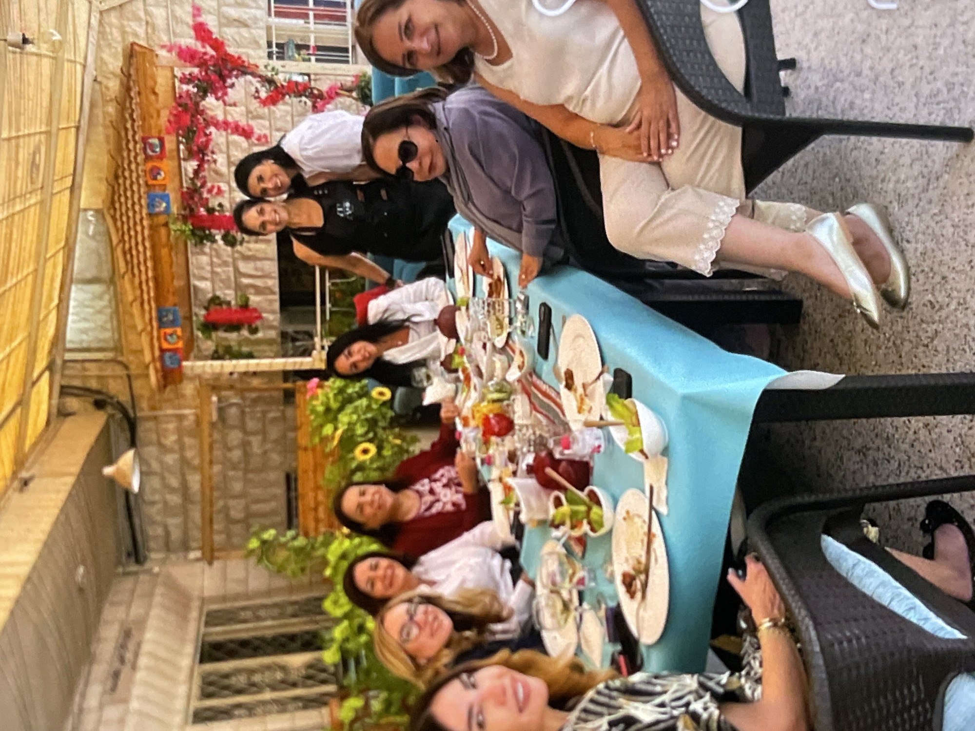 A group of nine women are seated around a long table covered with a bright blue tablecloth, enjoying a meal outdoors under a thatched roof. The table is laden with plates of food, glasses of wine, and various small dishes. The women appear to be in casual conversation and enjoying each other's company. The setting looks like a restaurant or a garden patio, with potted plants and decorative elements visible in the background. The overall atmosphere is relaxed and convivial.