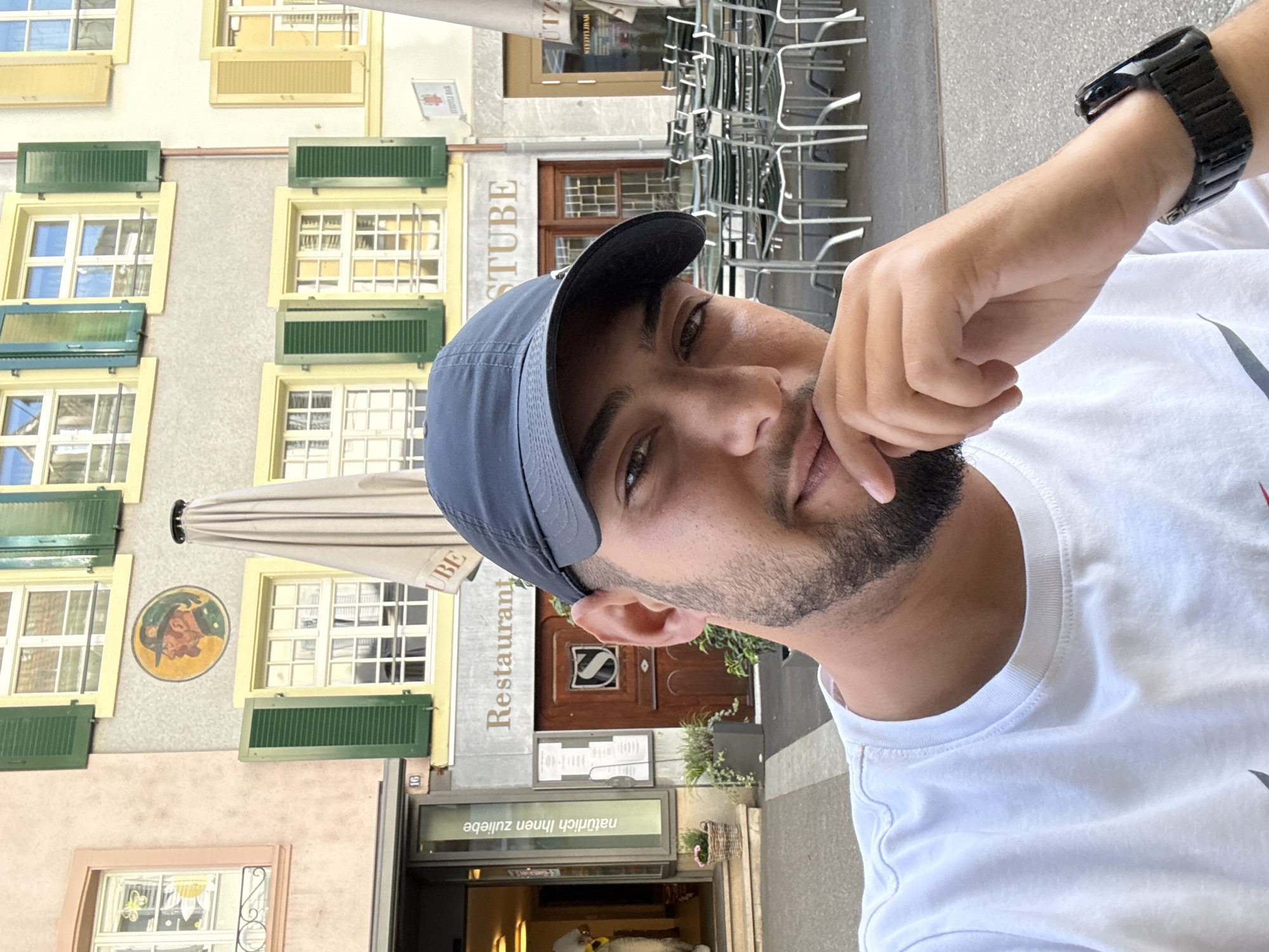 The image is a daytime selfie of a man outdoors, framed from the chest up. He has dark hair, a beard, and brown eyes, looking directly at the camera with a neutral to slightly contemplative expression. He wears a dark blue baseball cap and a white t-shirt, with a black-banded watch on his left wrist. His right hand rests near his mouth and chin.
The background reveals a traditional European building, likely a restaurant or inn, set in an urban environment consistent with Liestal, Switzerland. The multi-story structure features light-colored walls with numerous windows framed in yellow, many adorned with green shutters, some open and some closed. A round, colorful mural depicting a person's head in profile is painted on the building's facade.
Several signs are visible on the building. The prominent text "Restaurant" and "STUBE" identify the establishment. Below the main entrance, a glass panel reads "natürlich ihnen zuliebe," which is German for "naturally for your sake" or "naturally to please you." Above the "STUBE" sign, another sign states "SCHÜTZEN BAR." The number "16" is marked near a doorway. To the right, an outdoor seating area with numerous stacked metal chairs suggests a patio or café setting. The bright, clear lighting indicates it is daytime with pleasant weather.