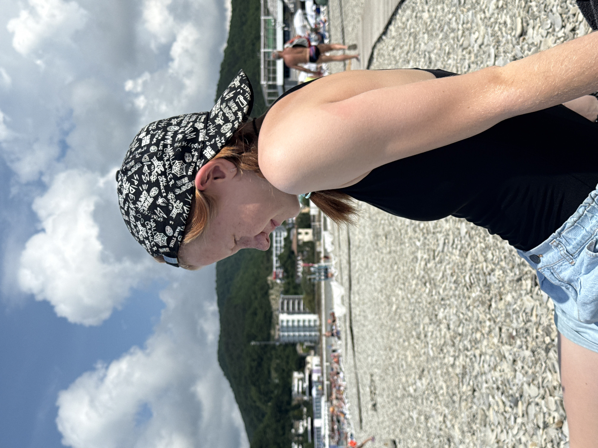 In Nebug, Russia, a young woman with red hair, wearing a black swimsuit, denim shorts, and a black baseball cap with a white pattern, sits on a pebble beach. Her face is turned to the side, and she is looking down. In the background, there are buildings, a green mountainside, other beachgoers, and umbrellas. The sky is blue with white clouds.