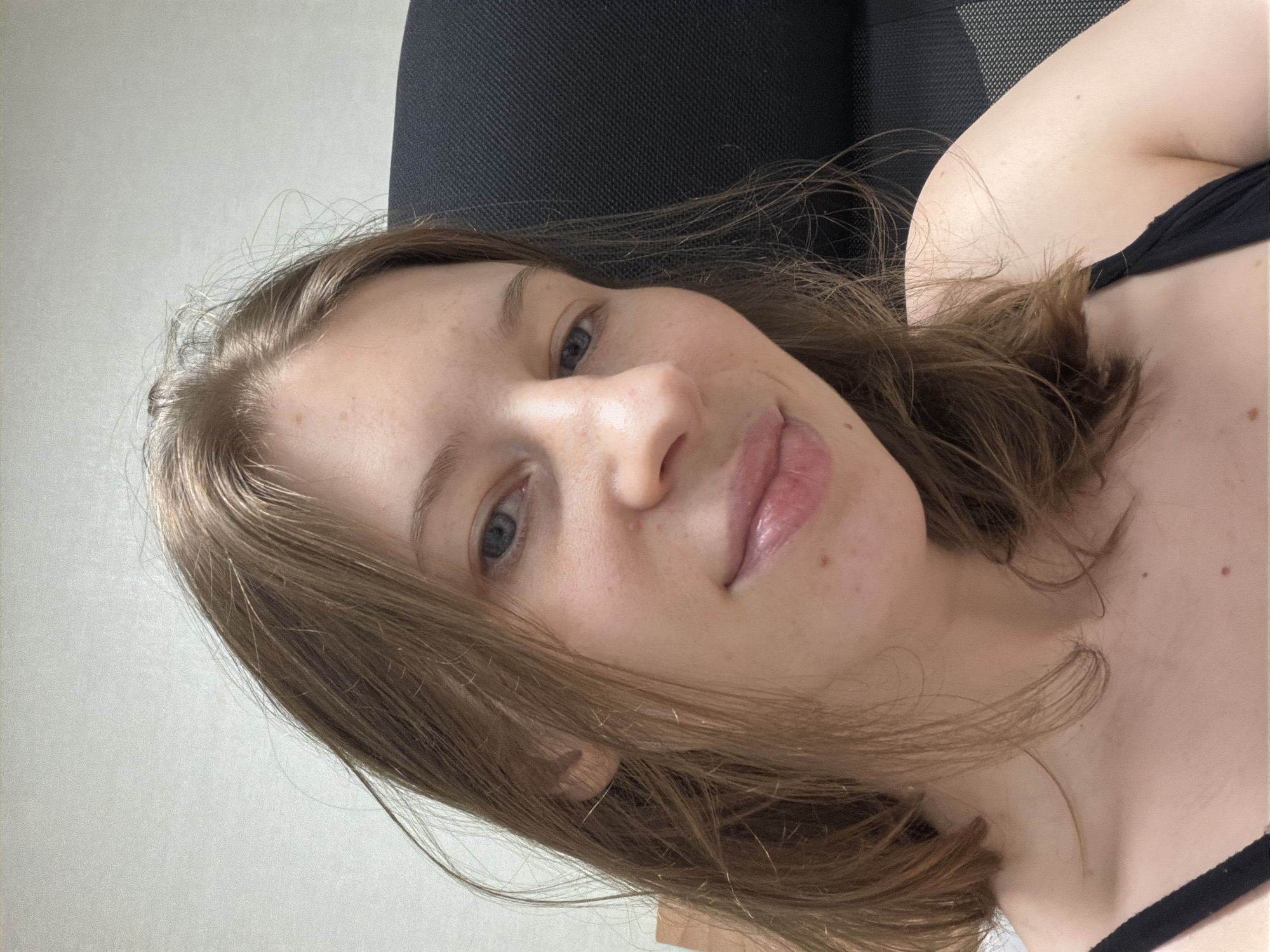 A young woman with light brown hair and blue eyes is shown in a close-up selfie. She is wearing a black tank top. Her skin has a few freckles. The background appears to be an indoor setting with a light-colored wall and a dark office chair. There is no indication of the specific location within Dongducheon-si, South Korea.