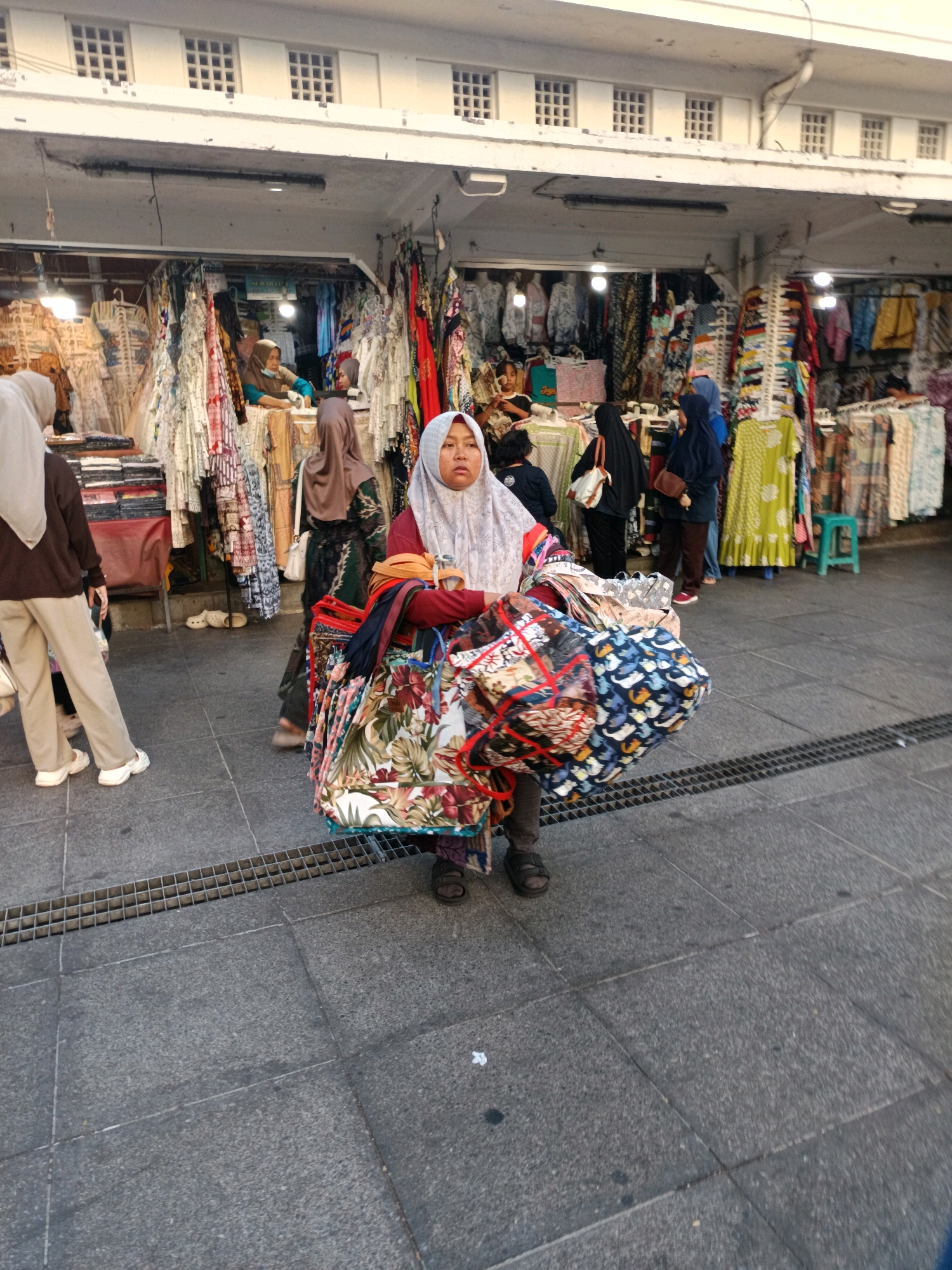 The image captures a bustling street scene in Yogyakarta, Indonesia, likely near a market or shopping area. In the foreground, a young woman wearing a light-colored hijab and a maroon top is carrying a large stack of fabric bags filled with textiles. Her expression appears somewhat strained or determined as she navigates the crowded walkway. To her left, another individual in a brown hijab and light beige pants walks away from the camera. Behind them, several market stalls are visible, displaying a colorful array of clothing and fabrics, with vendors attending to customers. The architecture of the buildings features a white facade with rows of square windows. The ground is paved with large, dark gray square tiles, and a metal grate runs along the walkway. The lighting suggests it is daytime, with sunlight casting shadows and illuminating the scene.