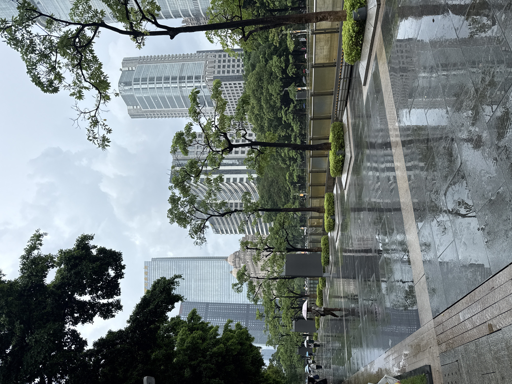 In Tianhe District, China, on a rainy day, a wet, paved pedestrian walkway reflects the surrounding modern skyscrapers and trees. Several people are walking on the path, some holding umbrellas. A person in the mid-ground is walking away from the camera, holding a light pink umbrella. Trees with lush green foliage are planted in rectangular beds along the walkway, with more dense foliage forming a backdrop of greenery behind them. The sky is overcast.