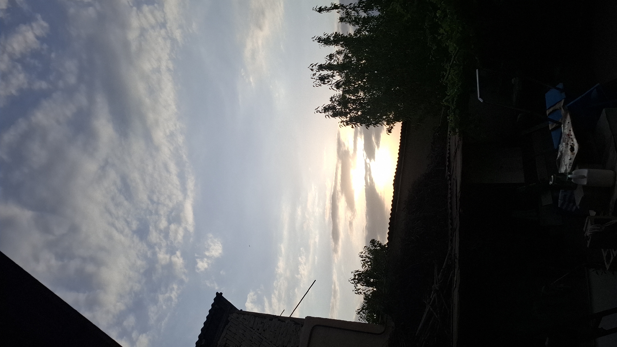 The image depicts a dusky sky over a residential area, likely in Fergana City, Uzbekistan. The sun is setting, casting a warm glow through scattered clouds. In the foreground, silhouetted against the sky, are rooftops and trees, with some architectural elements of buildings visible on the left. The lower part of the image is very dark, obscuring details of what appears to be an outdoor space with some objects, including what looks like a plastic jug and a table with items on it. The overall scene suggests the end of a day in a semi-urban or suburban environment.