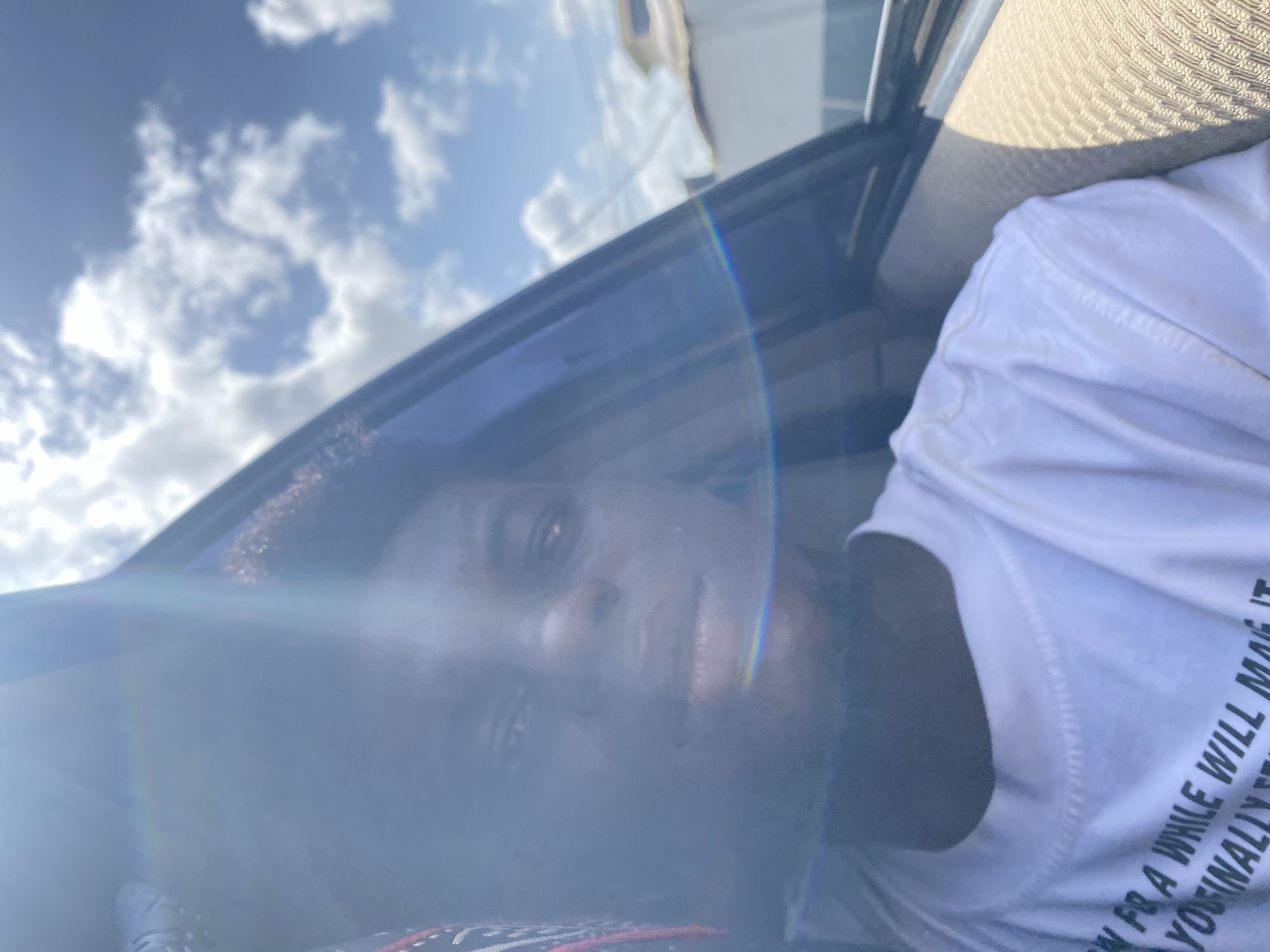 The image is oriented on its side, capturing an upward-angled view of a person inside a vehicle during the daytime. A prominent, rainbow-colored lens flare cuts diagonally across the frame from the bottom left to the top right, partially obscuring the subject's face and creating a bright halo effect.
The central subject is an individual with dark, curly hair, looking directly towards the camera. Their face is partially obscured by the intense sunlight and lens flare. They are wearing a white, short-sleeved t-shirt. Black text is partially visible on the front of the t-shirt, reading "...ON FR A WHILE WILL MAKE IT YOUINALLY".
Through a car window or sunroof, a clear blue sky with scattered white clouds is visible in the upper part of the image. Elements of the car's interior, such as a dark door frame and a light-colored, textured headliner or seat fabric, are discernible around the person, indicating the photo was taken from within a vehicle. The bright lighting and distinct shadows suggest it is a sunny day. The overall activity appears to be a selfie or a photograph taken from a low angle while inside the car.