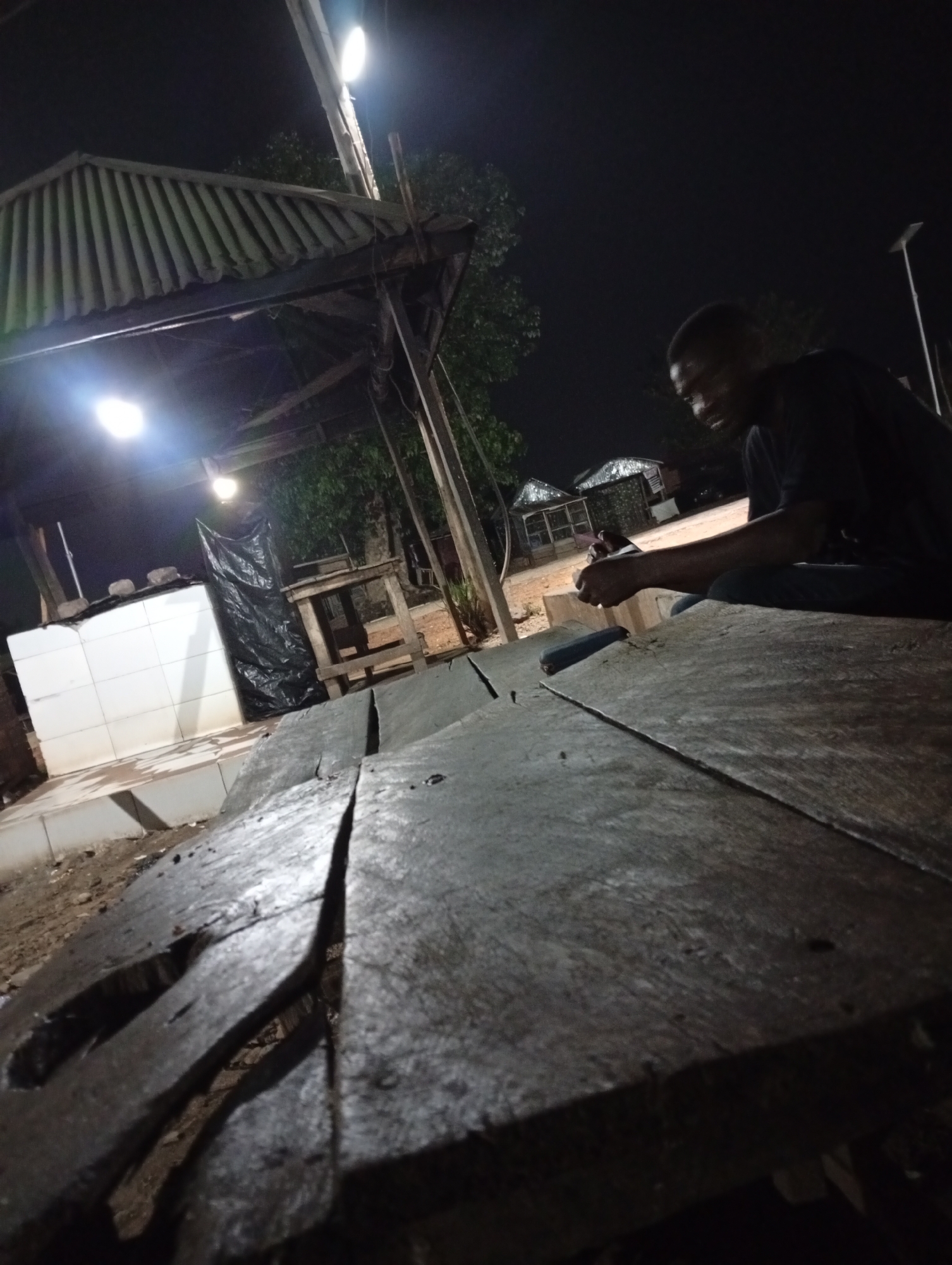 The image, taken at night in Adankolo, Nigeria, shows a man sitting on a weathered, rough-hewn wooden table outdoors. He is wearing a dark shirt and appears to be holding a small object in his hands, possibly looking at it. The table is made of thick planks with significant gaps and damage. Behind the table and to the left is a small, partially covered structure with white-tiled walls and a corrugated metal roof, illuminated by a bright light. Other structures, resembling small sheds or stalls, are visible in the background. The environment is dark, lit by streetlights and the light under the structure.