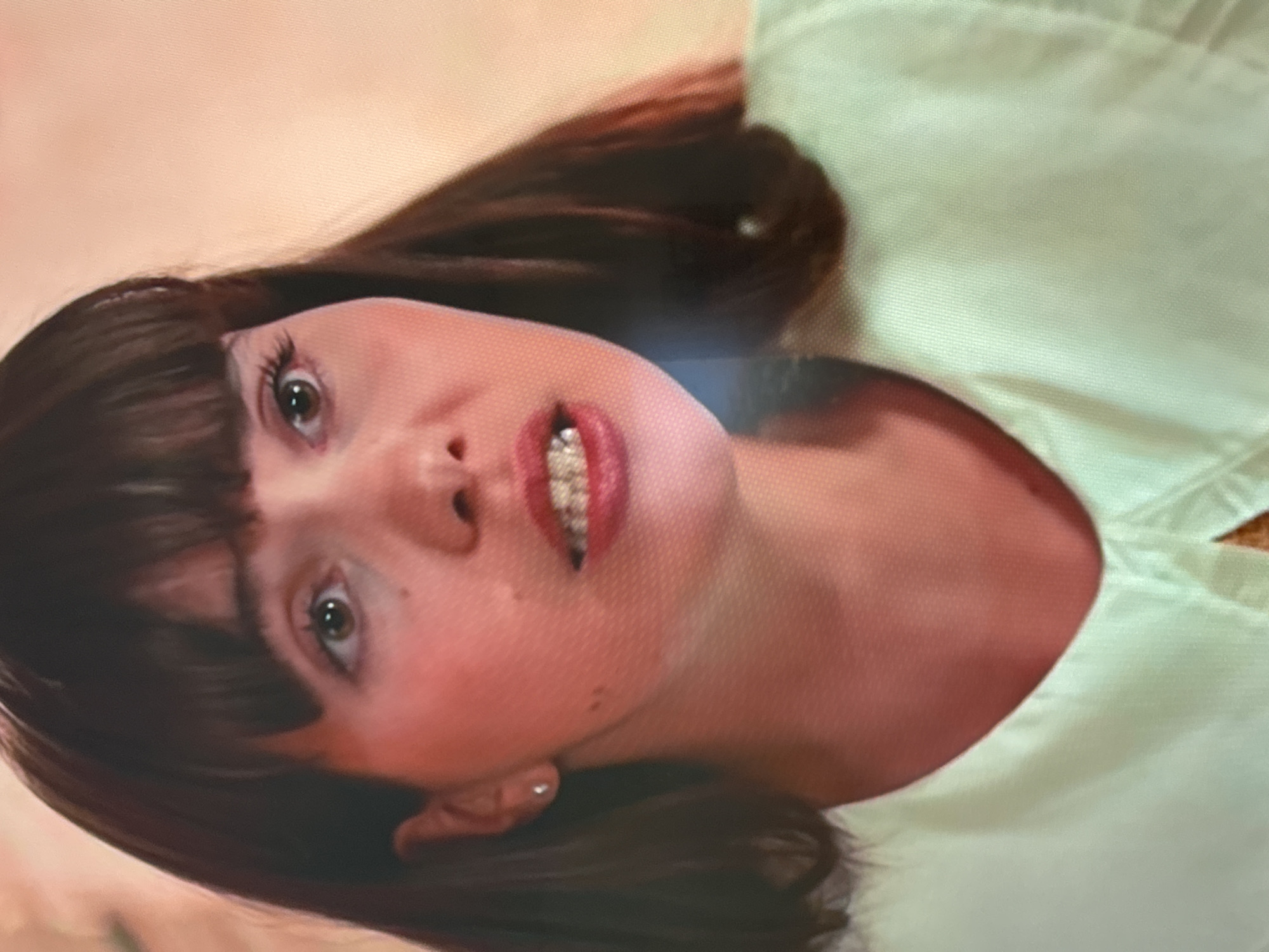 The image shows a close-up of a young woman's face and upper chest. She has dark brown hair with bangs, brown eyes, and full lips with red lipstick. She is wearing a light green collared shirt. She has a small stud earring in her left ear. The background is a soft, peachy-orange color. There is no indication of the city of Uyutnoye, Ukraine in the image.