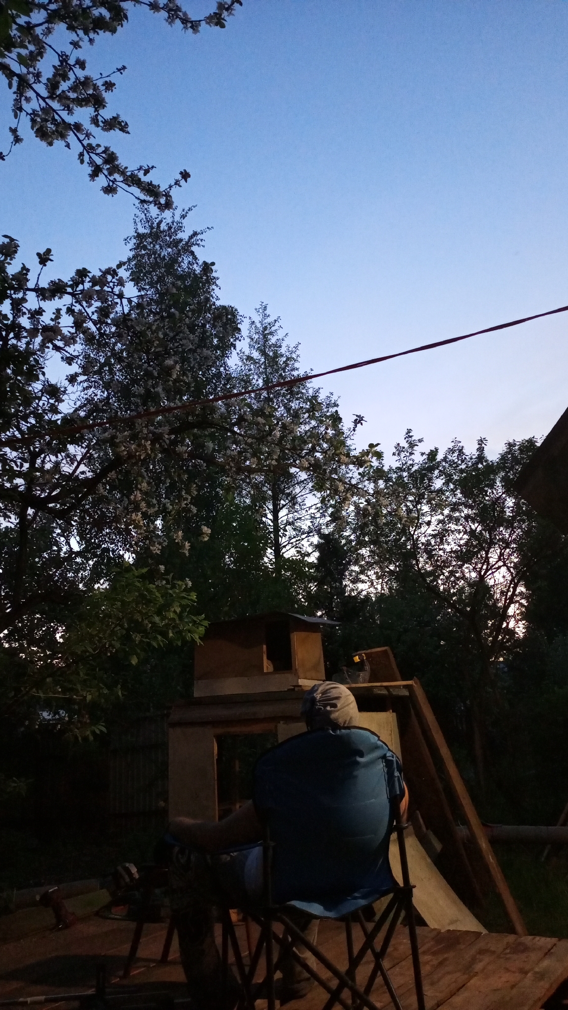 A person is sitting in a blue folding chair facing a partially constructed wooden structure in what appears to be a backyard in Юдино, Russia. The sky is a pale blue, transitioning to darker blue near the top of the frame. Trees with white blossoms are visible, framing the upper left corner of the scene. A red wire runs across the upper center of the frame. The area around the person is dimly lit.