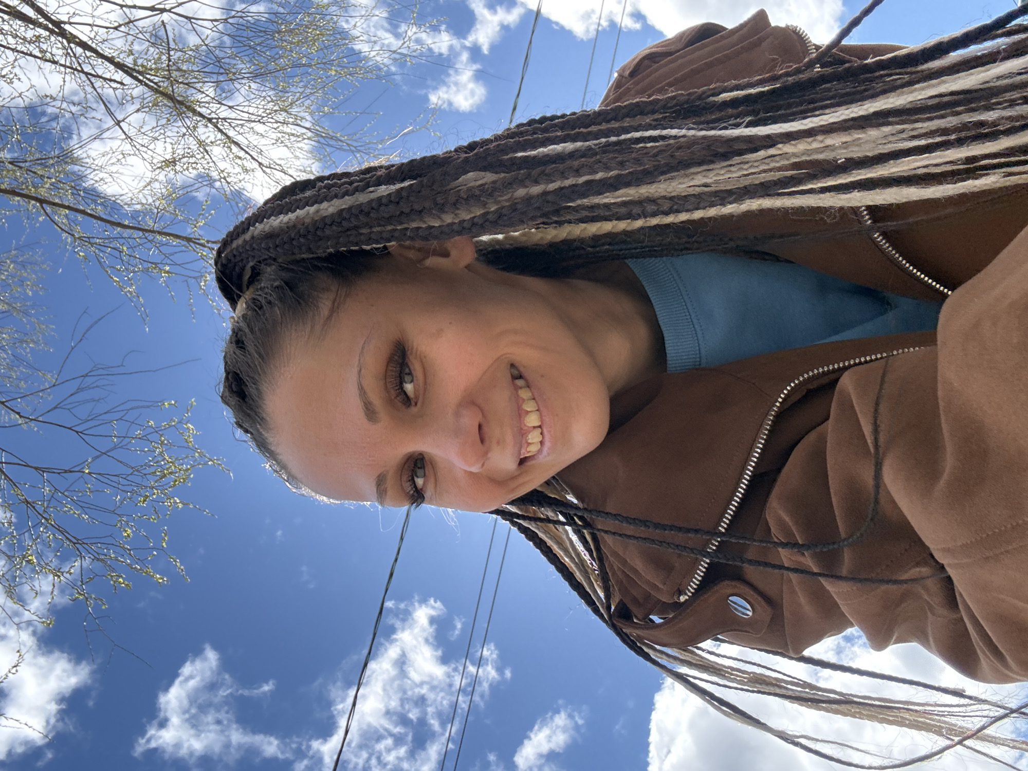 The image captures a young person, likely female, in a low-angle outdoor shot, looking downwards and smiling broadly at the camera. She has long, dark braided hair with distinct streaks of light brown/blonde and white/grey, which cascades over her shoulders. She is dressed in a light blue long-sleeved top under a brown jacket that features a visible silver zipper and a snap button detail.
The background is dominated by a bright blue sky dotted with scattered white, fluffy clouds, indicating clear and sunny weather. In the upper left corner, bare tree branches with small, light green buds suggest the season is early spring. Thin black power lines or wires stretch across the sky, hinting at an urban or suburban outdoor setting.
The subject's cheerful smile conveys a positive emotion. The bright daylight suggests the photo was taken during the daytime. This scene is set in Rusanovo, Russia. There is no visible text in the image.