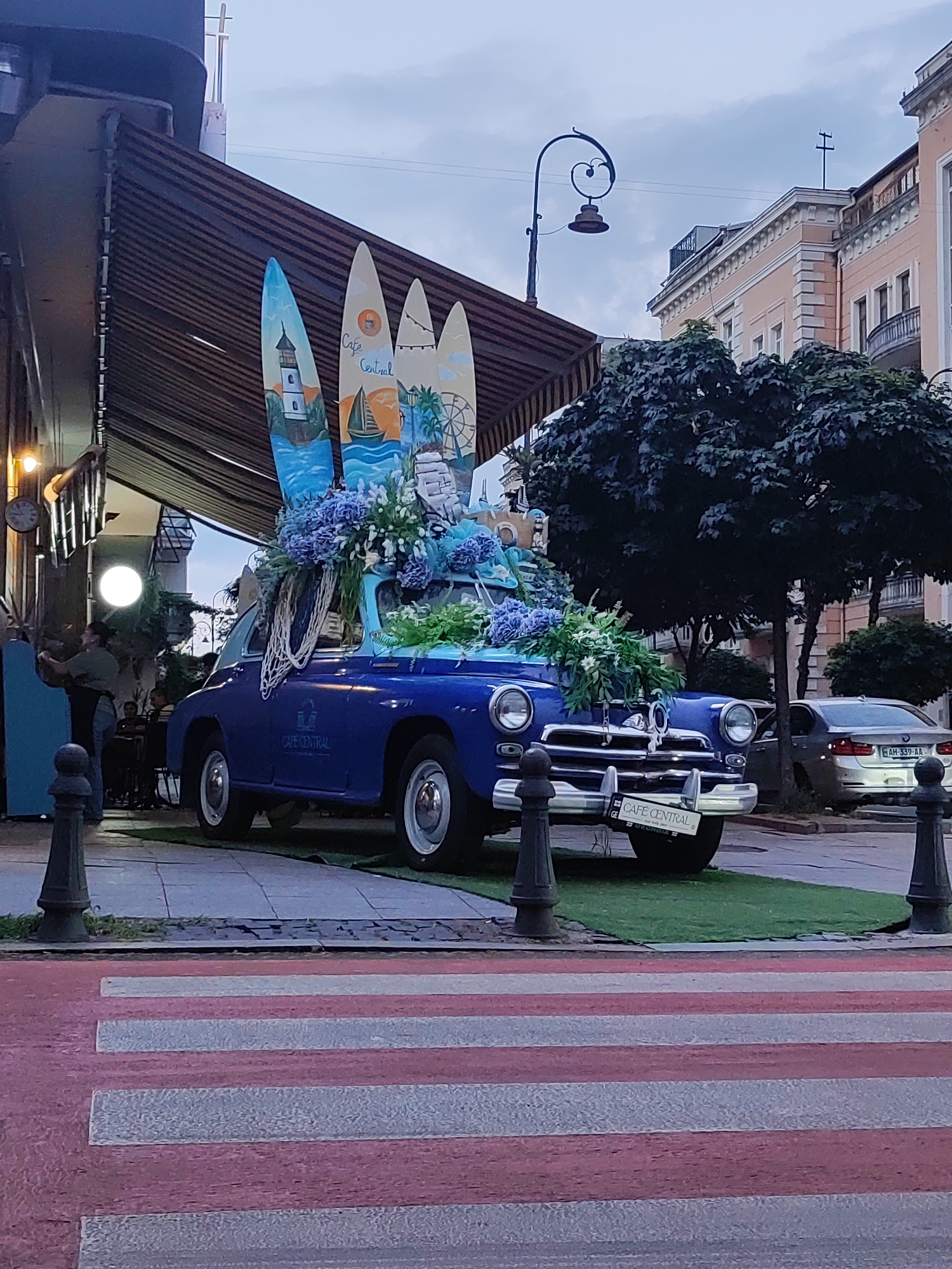 The image depicts a blue vintage car, likely a GAZ-21 Volga, adorned with numerous surfboards and floral arrangements. The car is positioned on a patch of artificial grass in front of a building with an awning. The surfboards are decorated with painted scenes of lighthouses, sailing ships, and a Ferris wheel. The car has a license plate that reads "CAFE CENTRAL" and "GEORGIA". In the background, there are buildings with classical architecture, trees, and a modern silver car. A crosswalk with red and white stripes is visible in the foreground. The overall scene suggests an outdoor cafe or promotional display, possibly in Batumi, Georgia, given the license plate information and architectural style. The sky is overcast.