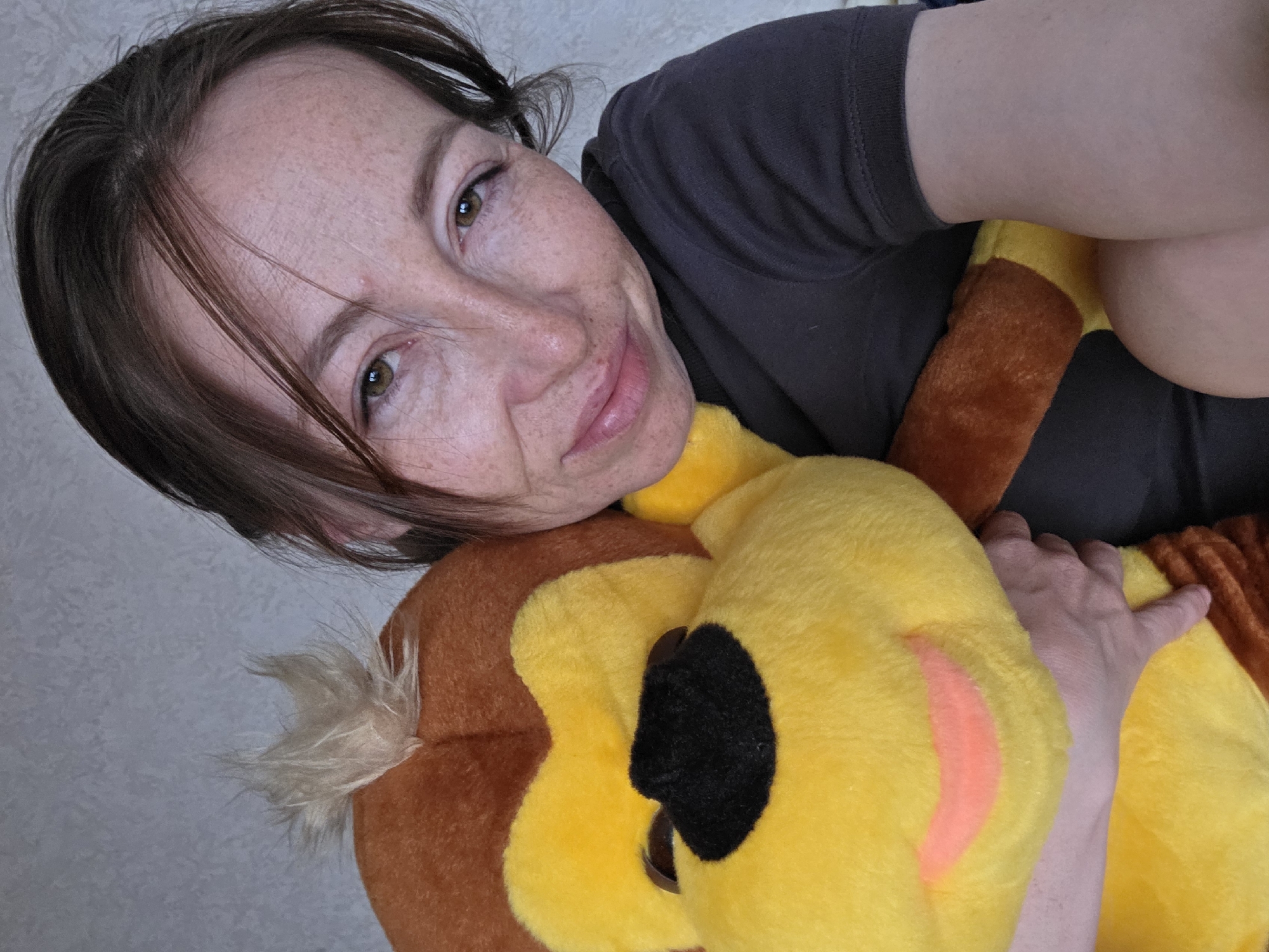 A woman with brown hair and freckles is holding a large, plush lion toy with yellow and brown fur. She is wearing a gray t-shirt. The background is a plain, light-colored wall. The location is Unknown, Unknown.