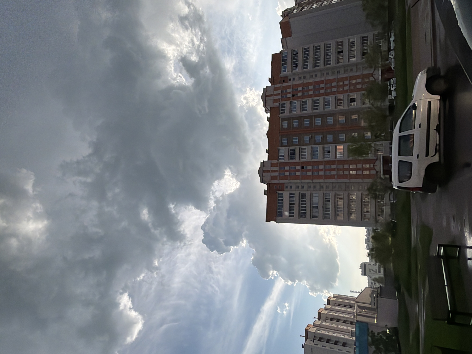 The image captures a scene in what appears to be an urban residential area. A tall, multi-story building with a red and white facade dominates the view, juxtaposed against a cloudy sky. A white van is parked on the right side of the frame along a paved street lined with greenery. In the distance, there are glimpses of other buildings and infrastructure. The overall atmosphere suggests an overcast day in an urban environment.