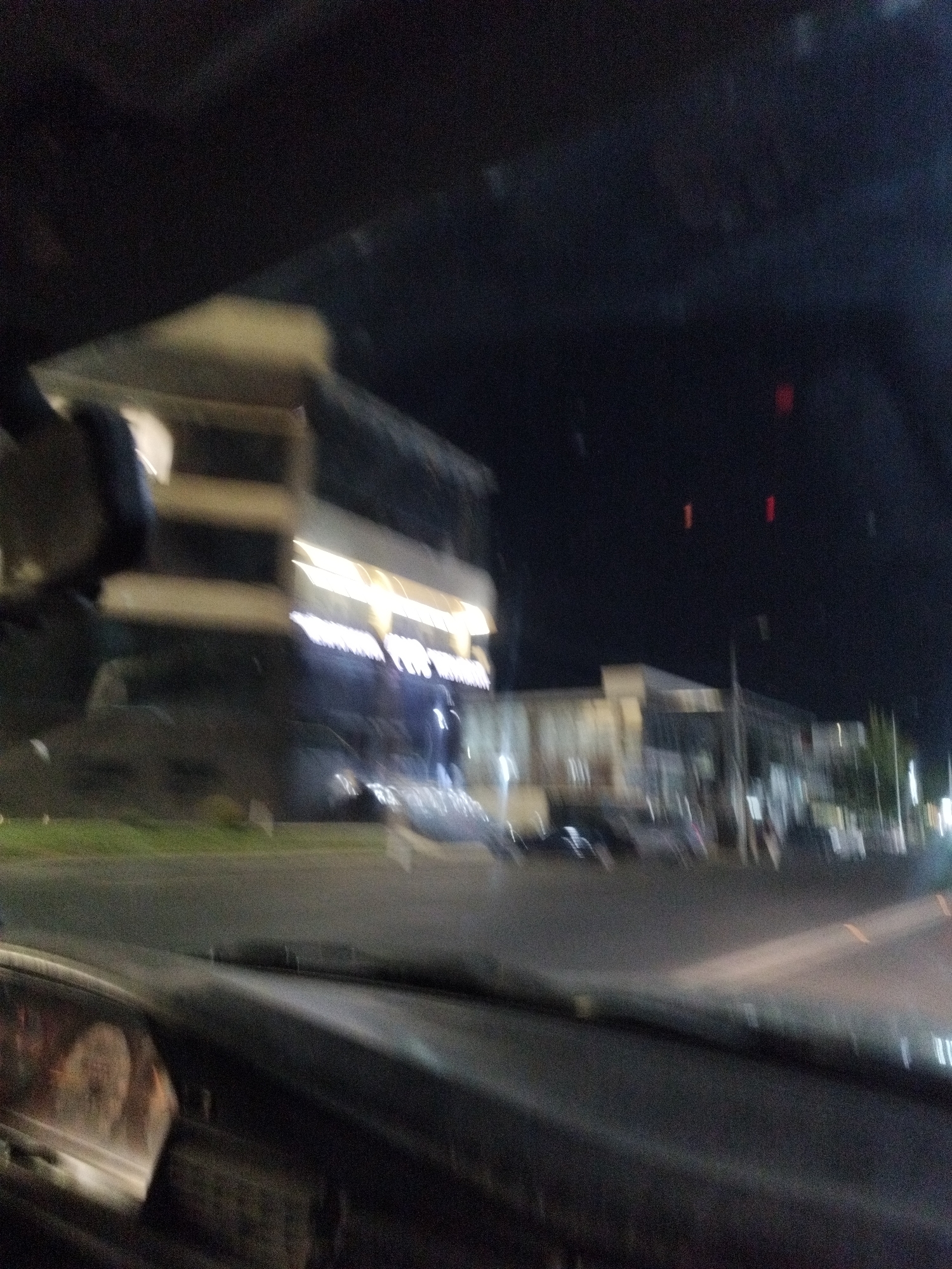 The image, taken from inside a vehicle at night, shows a blurry view of a street scene in Shymkent, Kazakhstan. A multi-story building with illuminated windows is visible on the left, featuring horizontal bands of light. Across the street, other buildings and streetlights are present, but they are out of focus. The ground appears to be asphalt, and there is a patch of green grass on the left side of the road. The interior of the vehicle, including the dashboard and what appears to be a reflection on the windshield, is also visible in the foreground. The overall impression is of a moving vehicle capturing a nighttime urban environment.