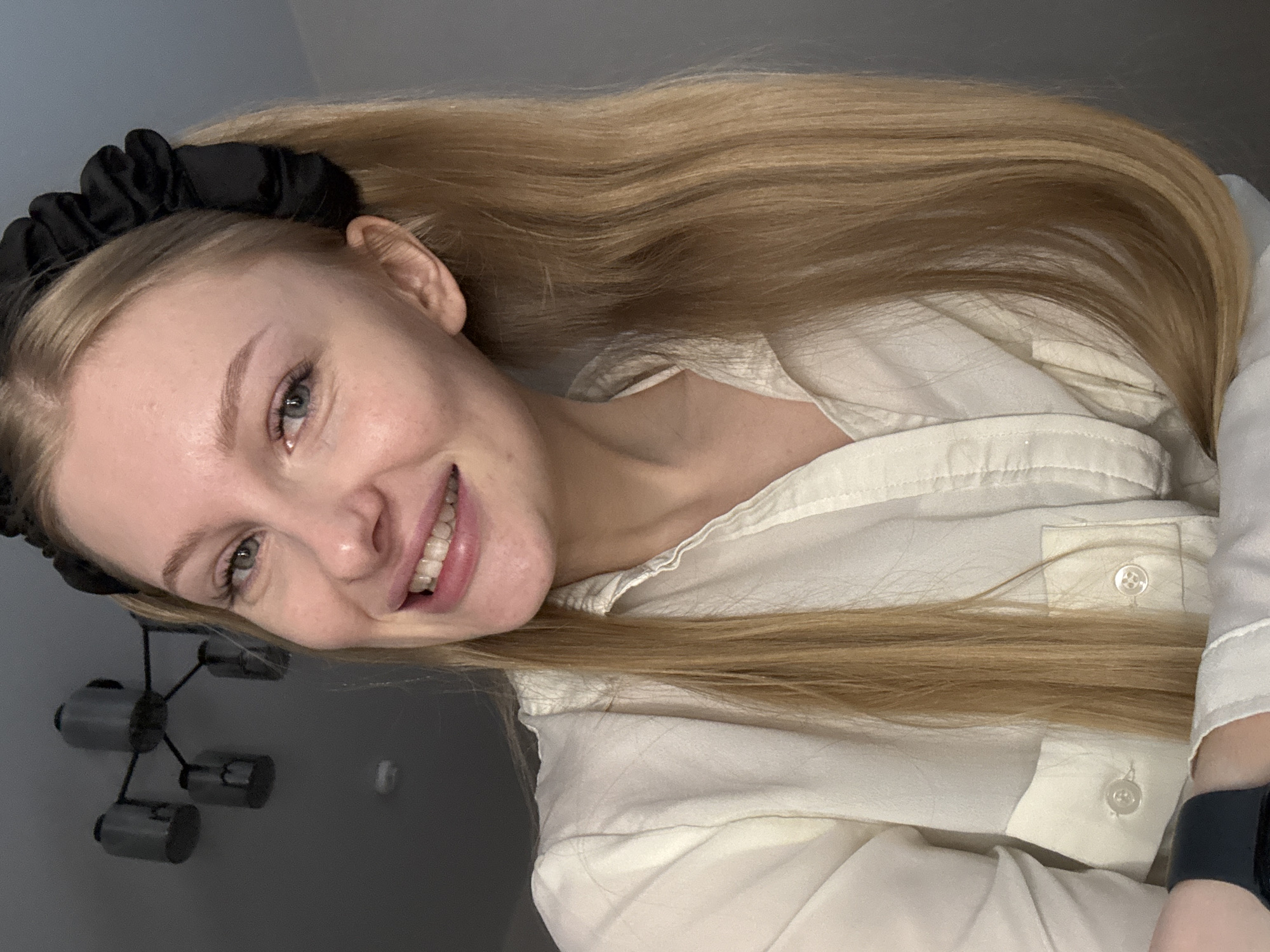 A young woman with blonde hair tied in pigtails with a black scrunchie smiles at the camera. She is wearing a white button-down shirt. The background is a plain gray wall, with a black chandelier hanging from a dark ceiling in the upper left corner. There is no discernible location in the city of Perm, Russia.