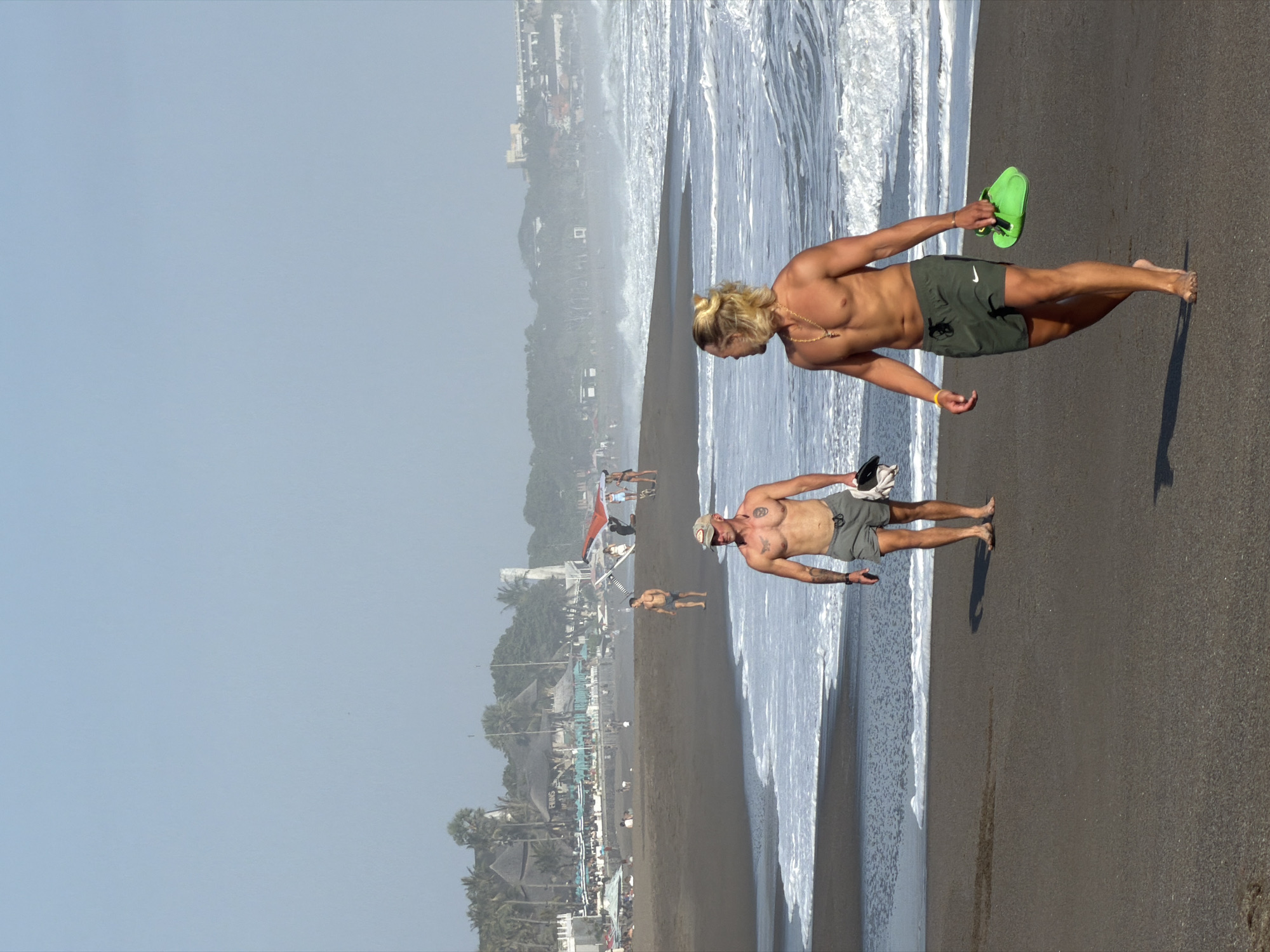 The image depicts a beach scene in North Kuta, Indonesia, with two men walking on the dark sand. The man in the foreground, on the right, has blonde hair pulled back and is wearing green swim shorts. He is holding a pair of bright green flip-flops. The second man, further back and to the left, is wearing gray swim shorts and has tattoos visible on his chest and arm. He is also holding a dark object, possibly a towel, in his hand. The ocean with white waves is to the right of the men, and the background shows a hazy shoreline with palm trees and buildings. Other beachgoers and structures are visible in the distance. The sky is clear and pale blue.